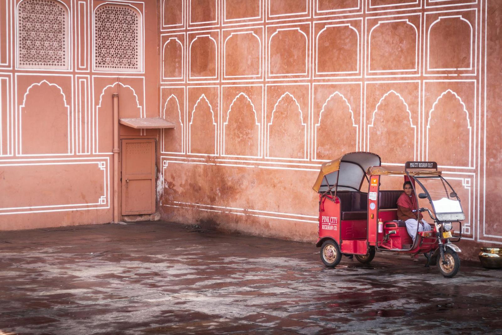 A red auto-rickshaw parked against a pink wall painted with white archway patterns