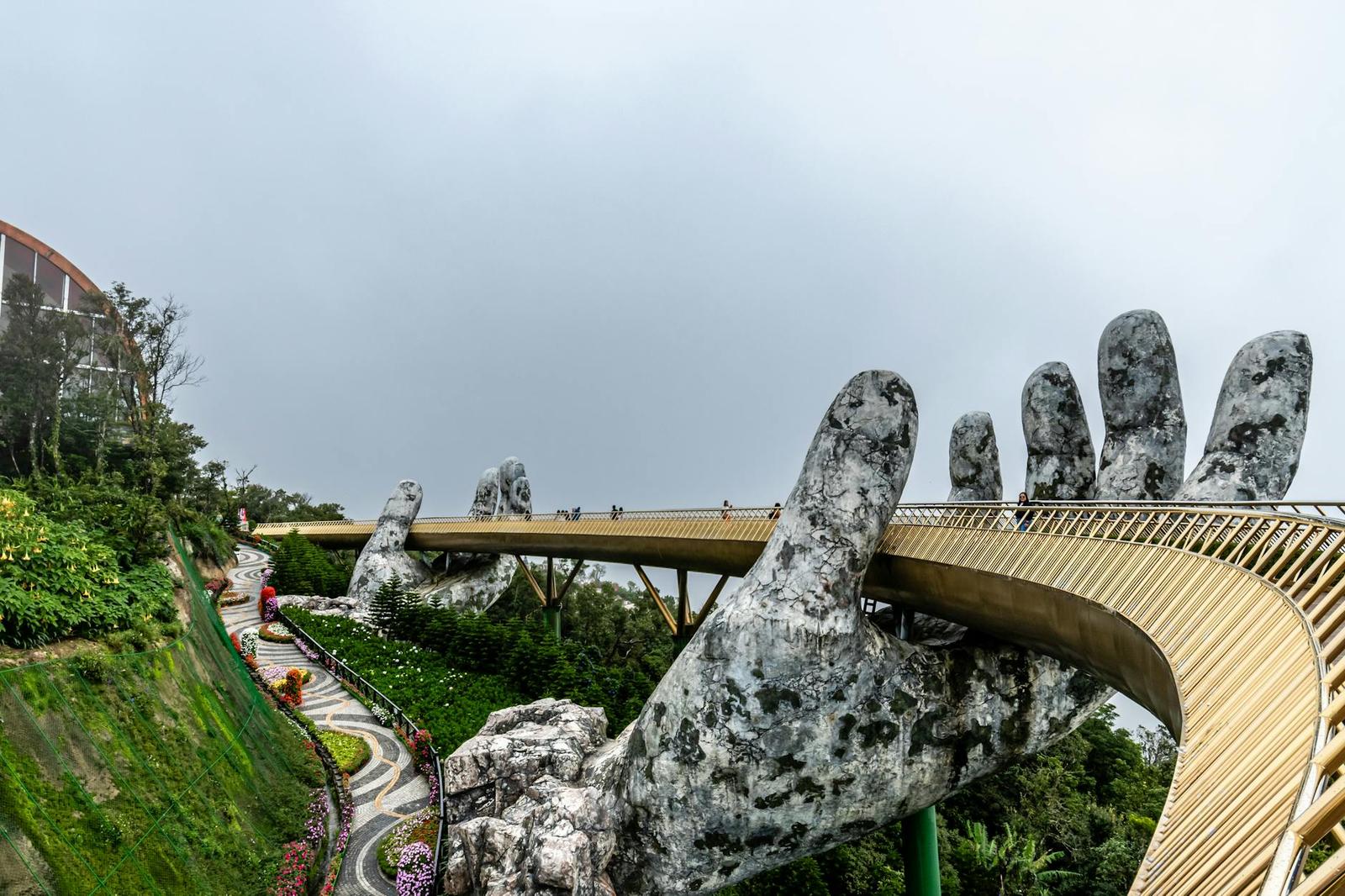 A curved golden bridge held by giant stone hands winding down a landscaped hillside