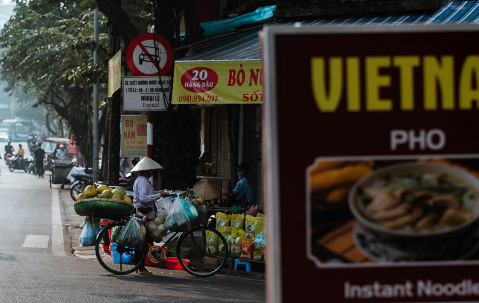 Street vendor with conical hat and loaded bicycle beside a noodle shop sign