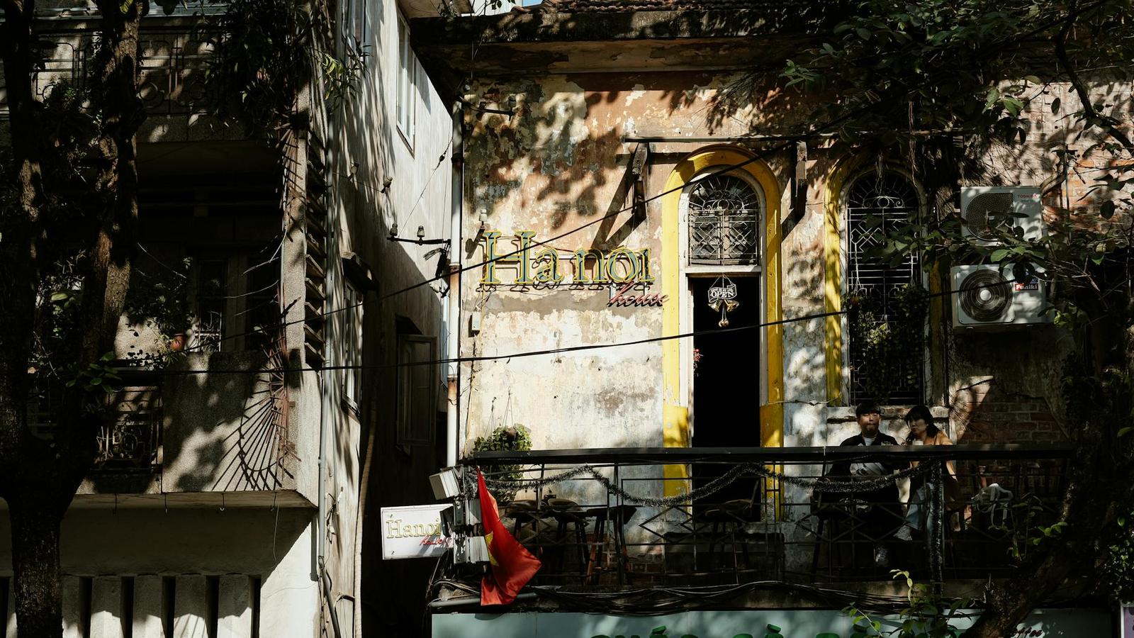 A weathered colonial building facade with a balcony sign and a person seated behind shutters