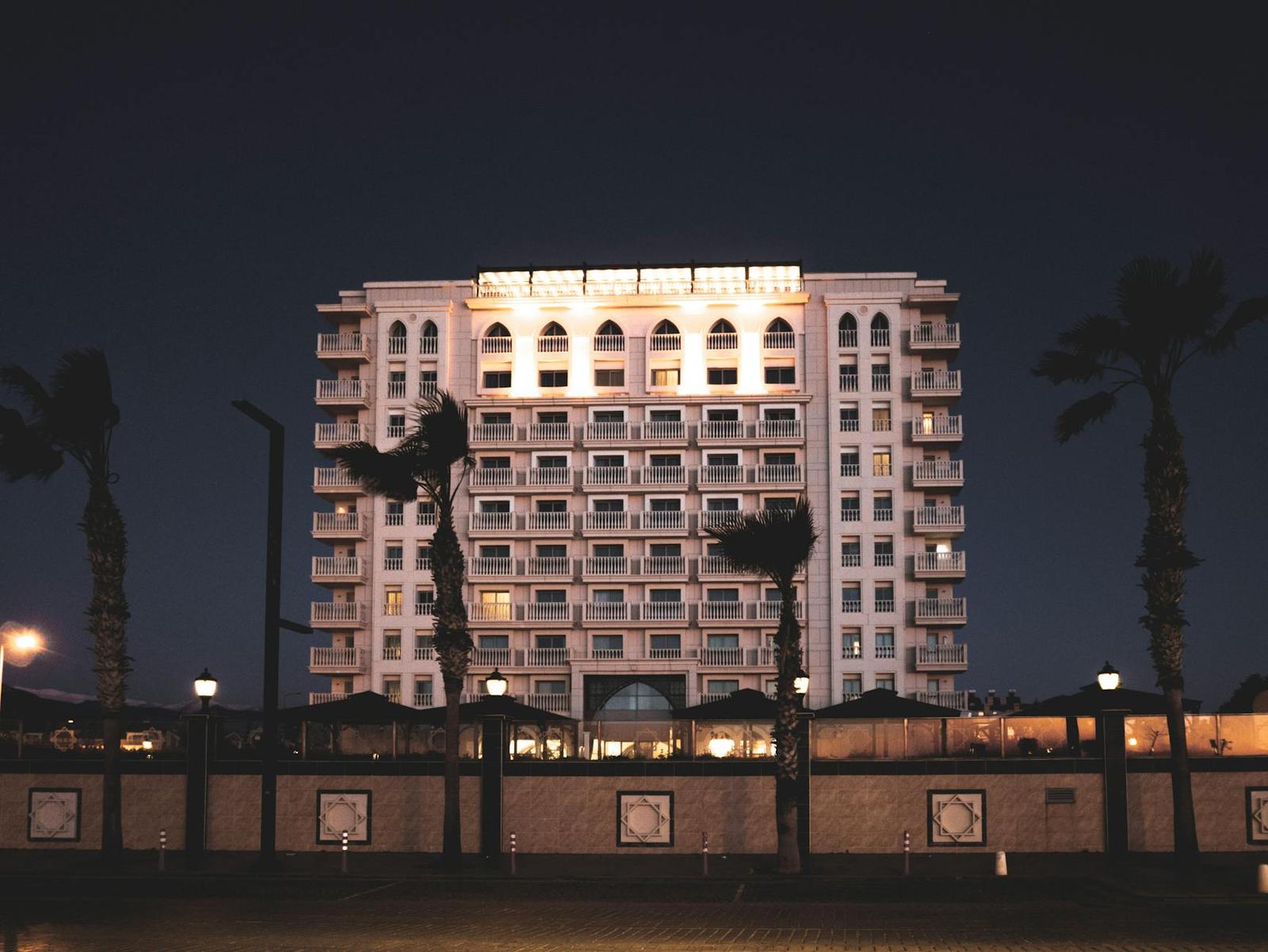 A tall hotel building lit up at night behind palm trees and a wall