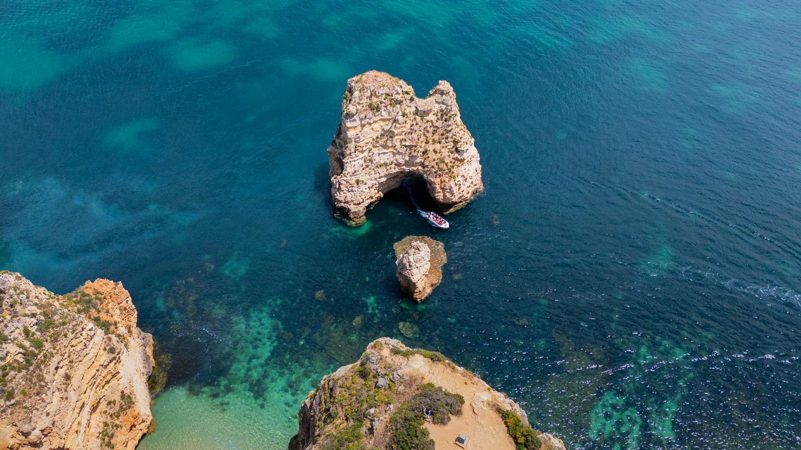 A small boat passes a weathered rock arch rising from clear turquoise water