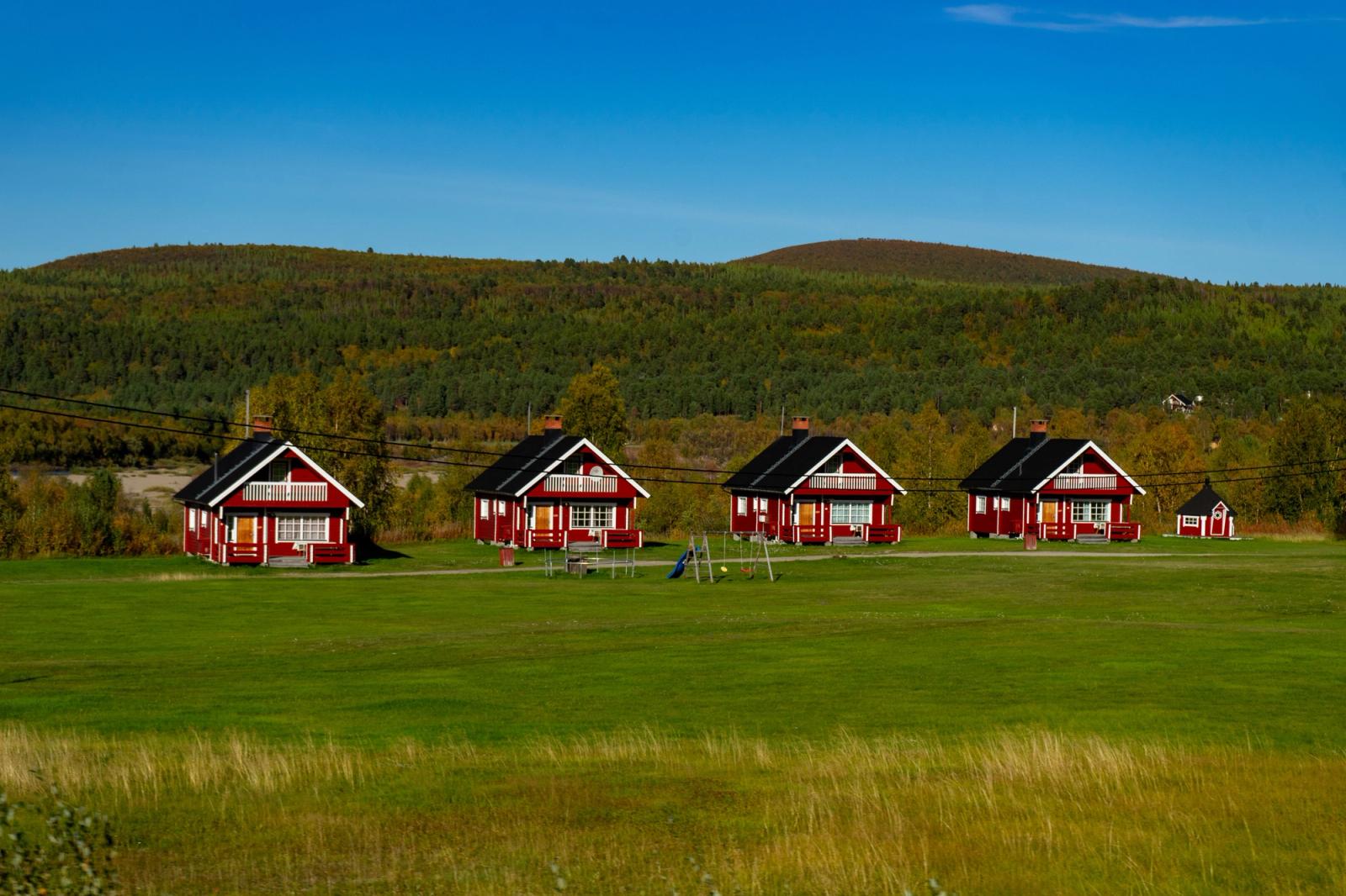 Red wooden cabins in Norway's rural landscape surrounded by lush greenery and hills under a clear blue sky