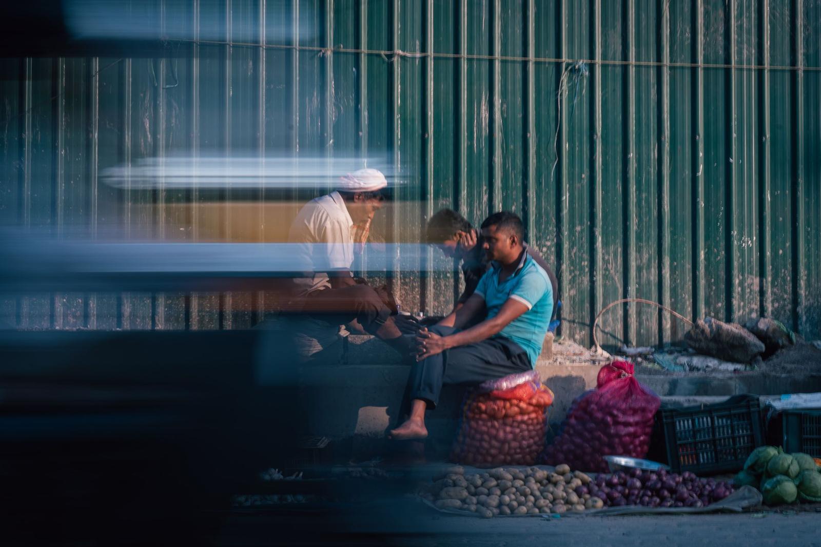 Two men sit by roadside sacks of produce as traffic blurs past