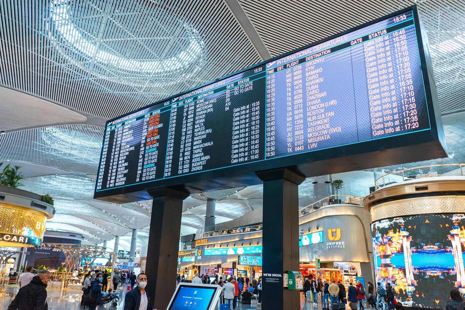 Travellers pass beneath a large flight information board in an airport terminal