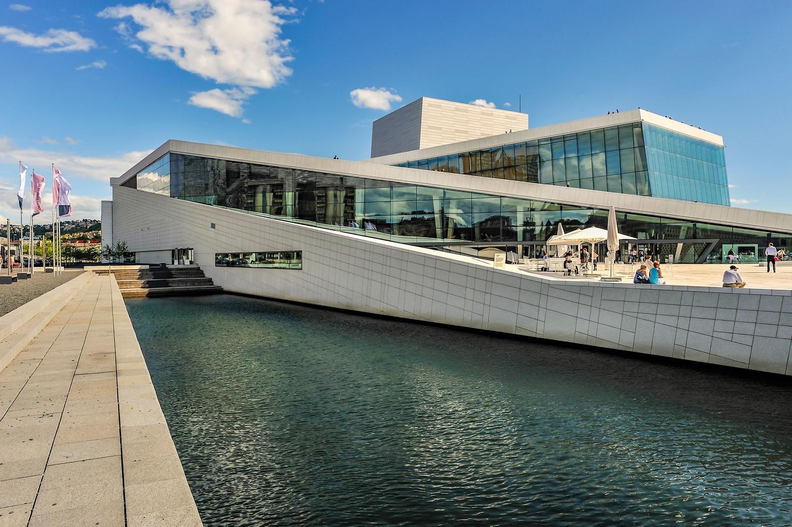An angular white building with sloped roof terraces beside a still waterfront canal