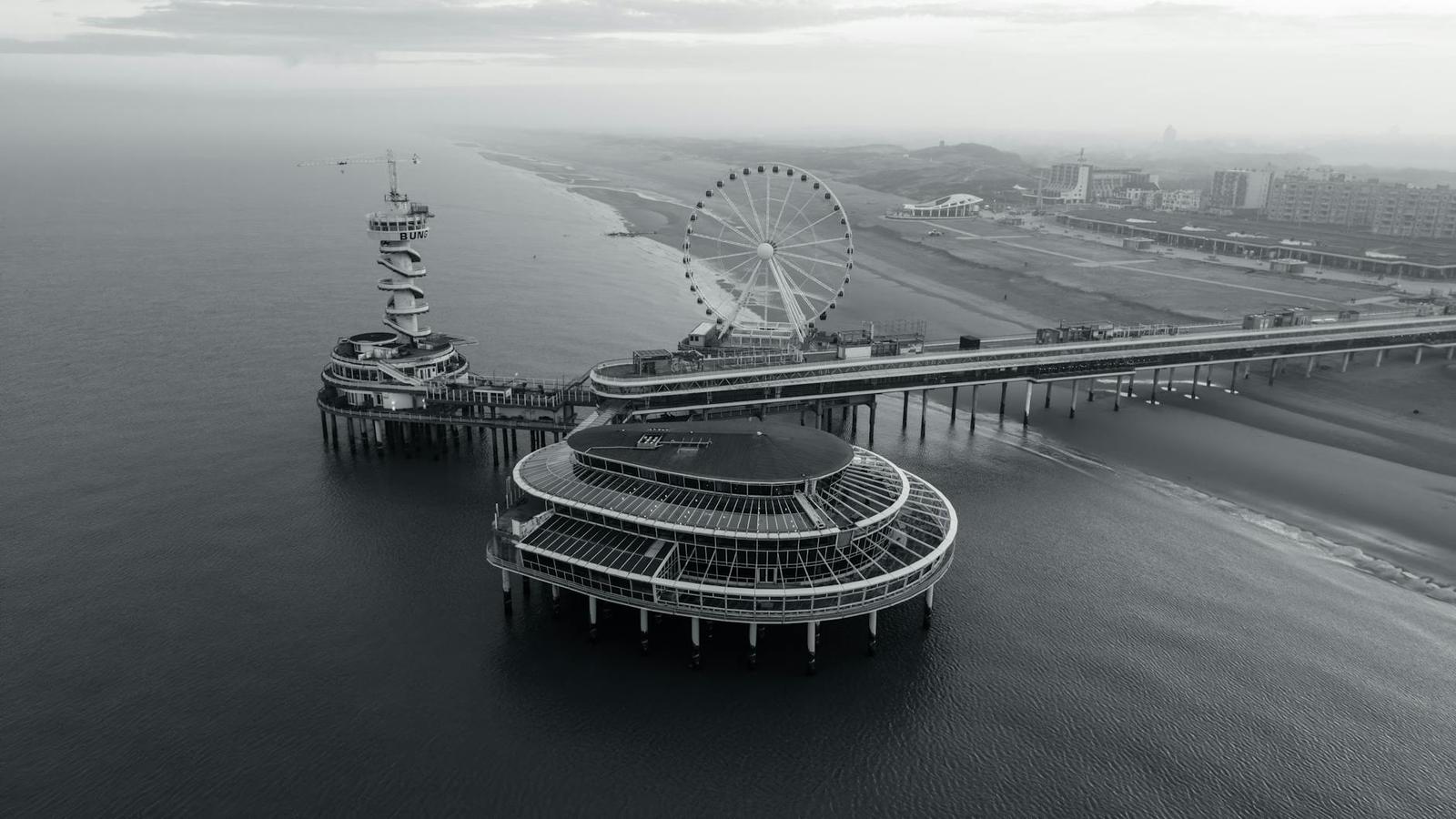 An aerial monochrome view of a pier with a ferris wheel over the sea