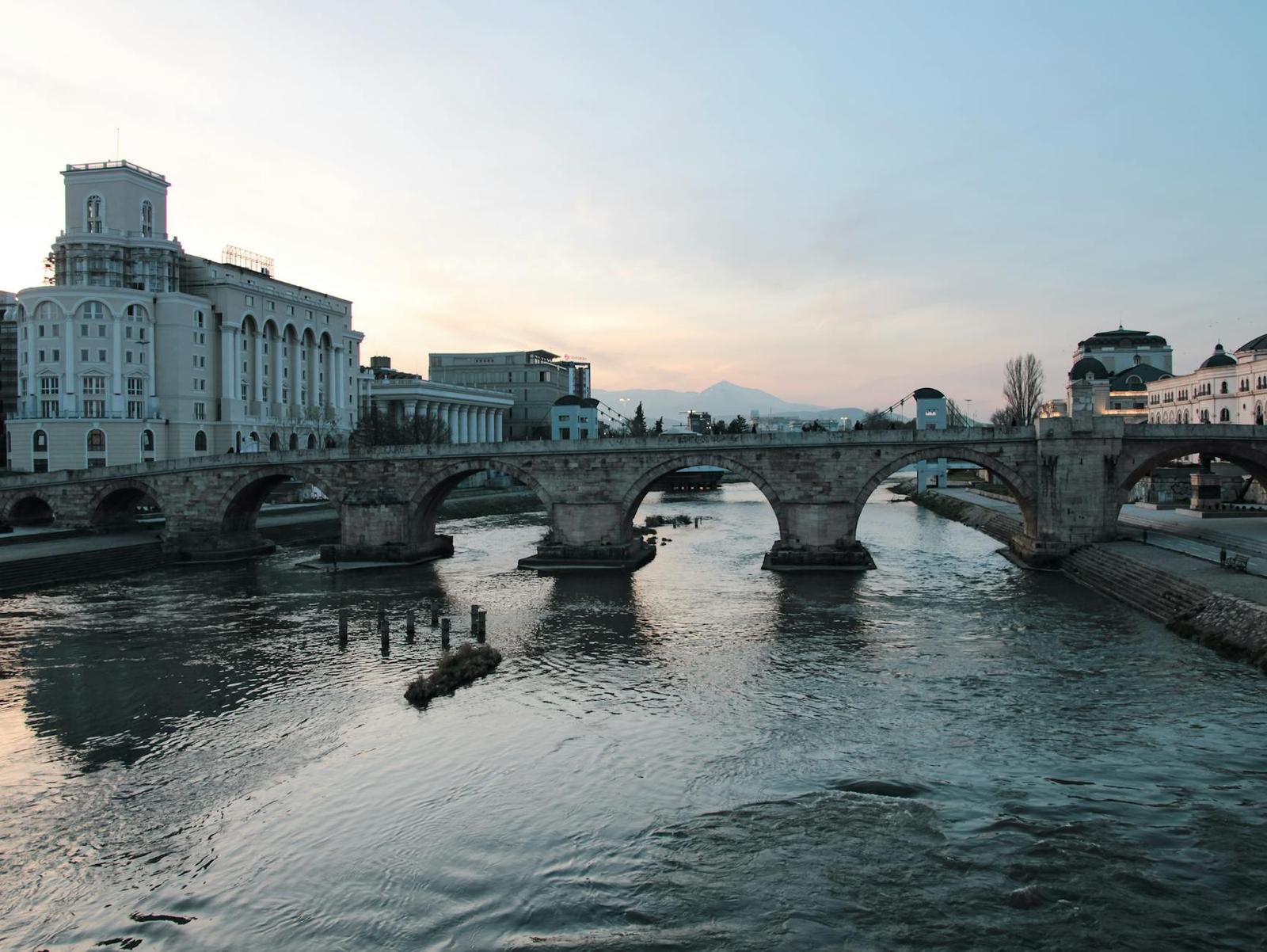 A stone arched bridge crossing a river beside a city skyline at dusk