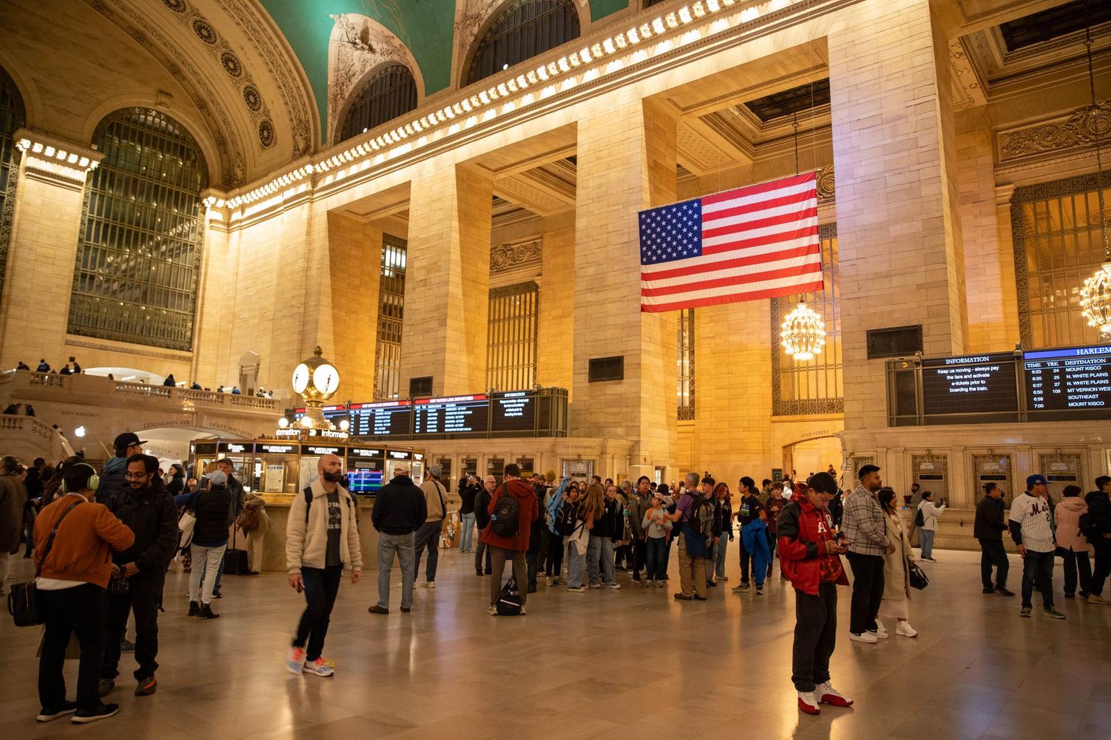 Travelers crossing a grand marble hall beneath a large hanging flag and departure boards