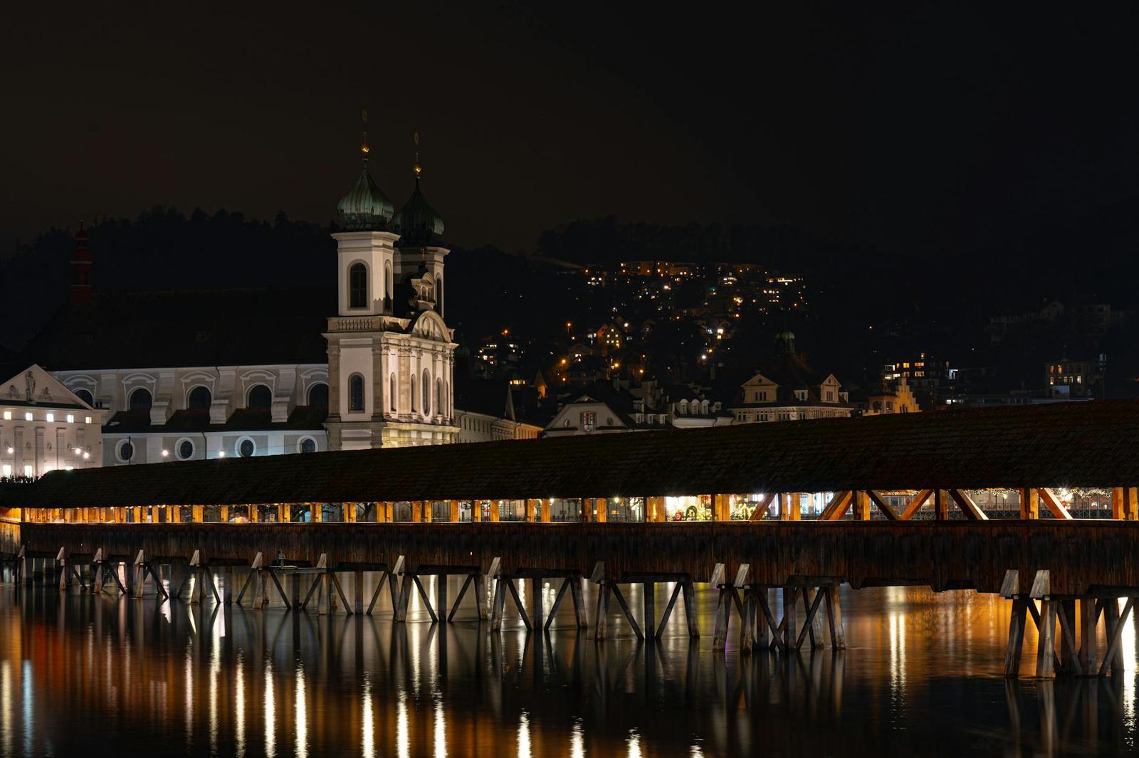 Illuminated church towers and a covered wooden bridge reflected in a river at night