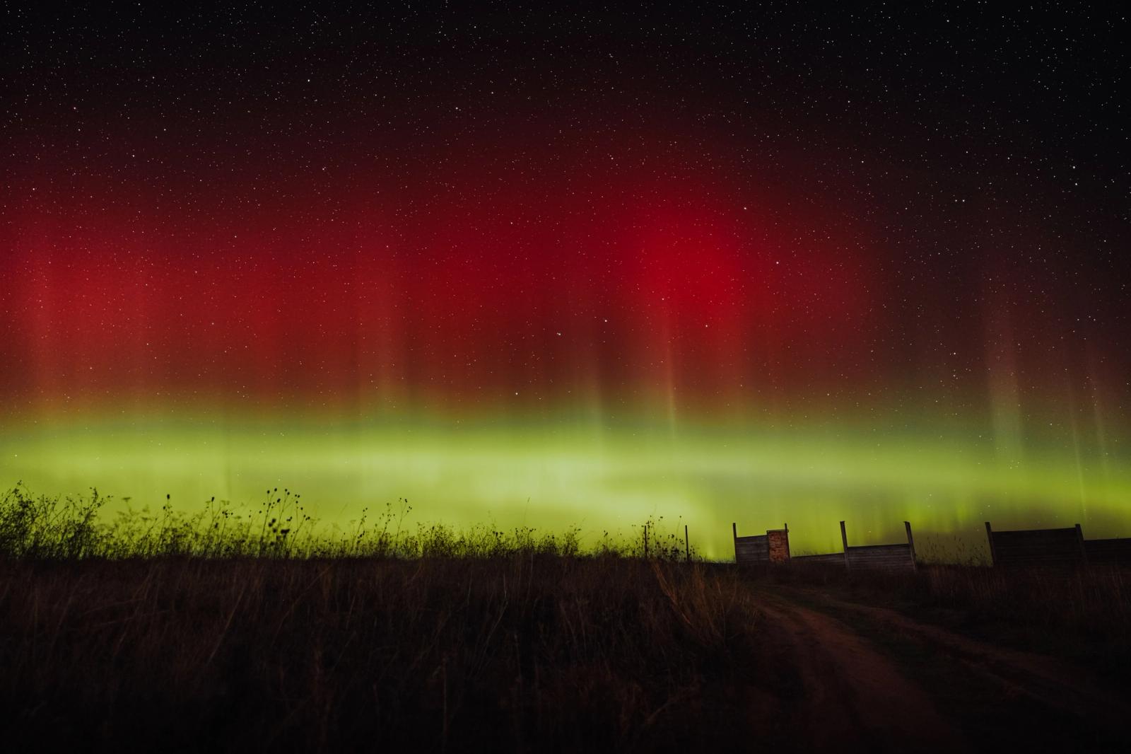 Red and green aurora pillars over dark farmland with silhouetted rooftops