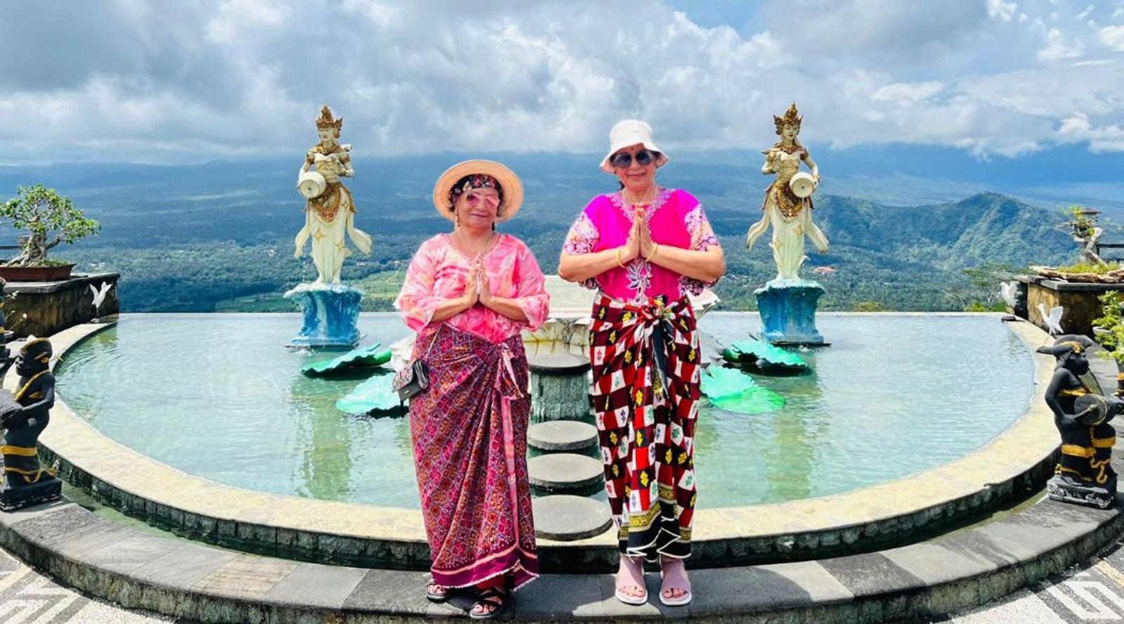 Two women in traditional attire posing by a scenic fountain.