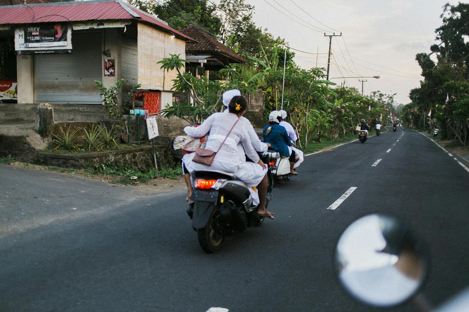 Riders in white ceremonial dress on scooters along a rural road