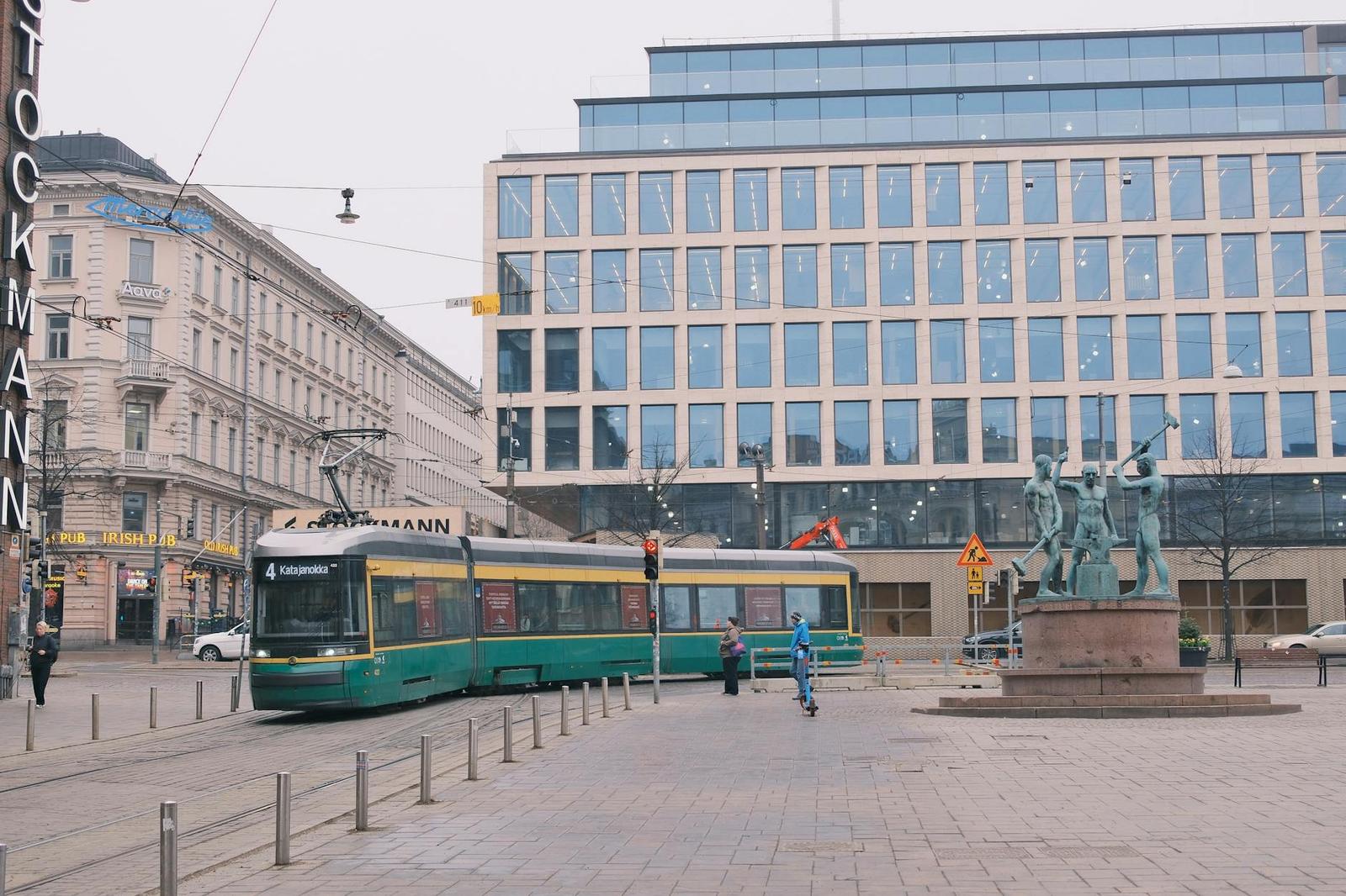 A green tram passes a glass office building and public statue on a city square