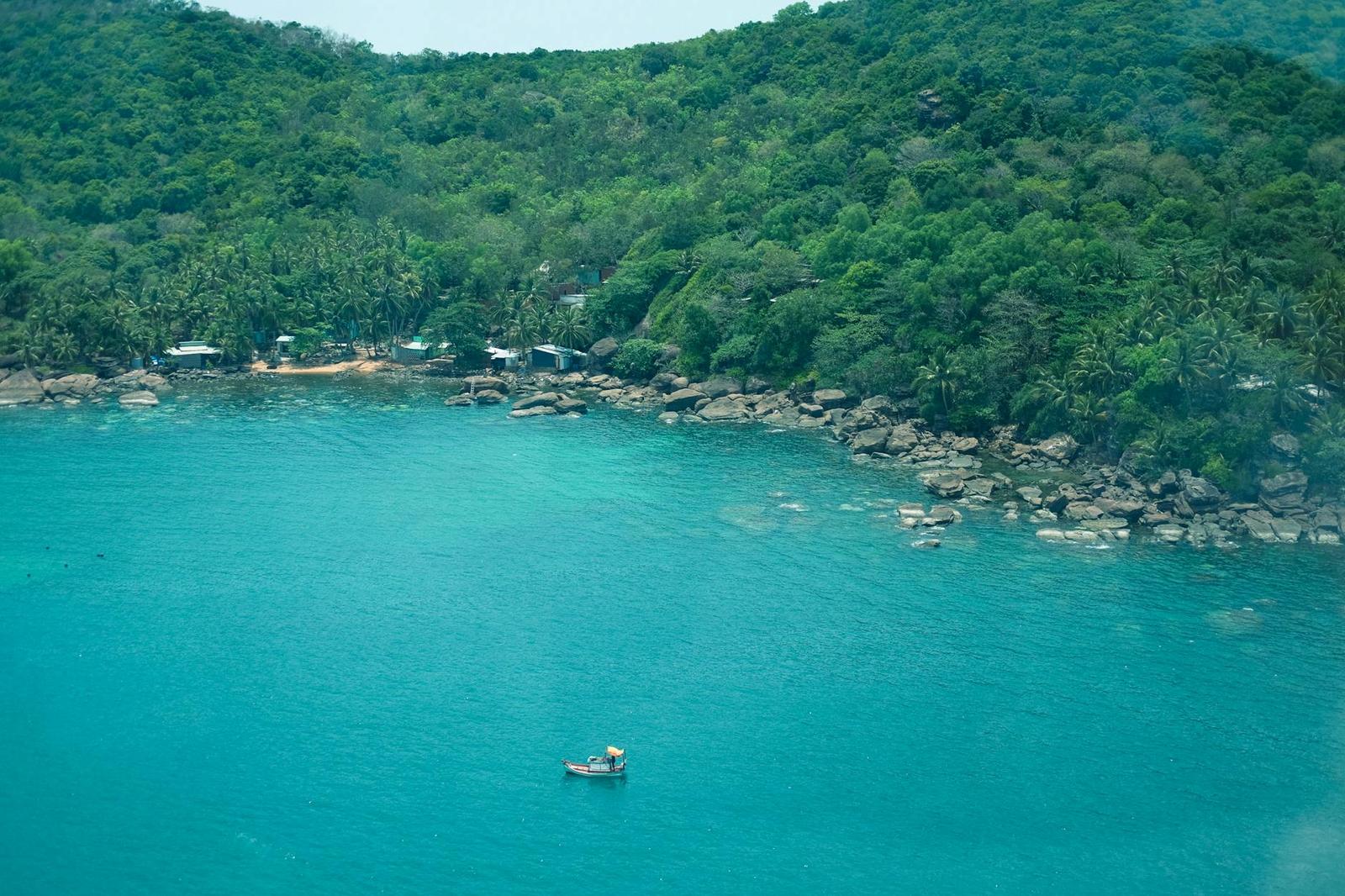 Aerial view of a small boat on a turquoise bay below a forested rocky coastline