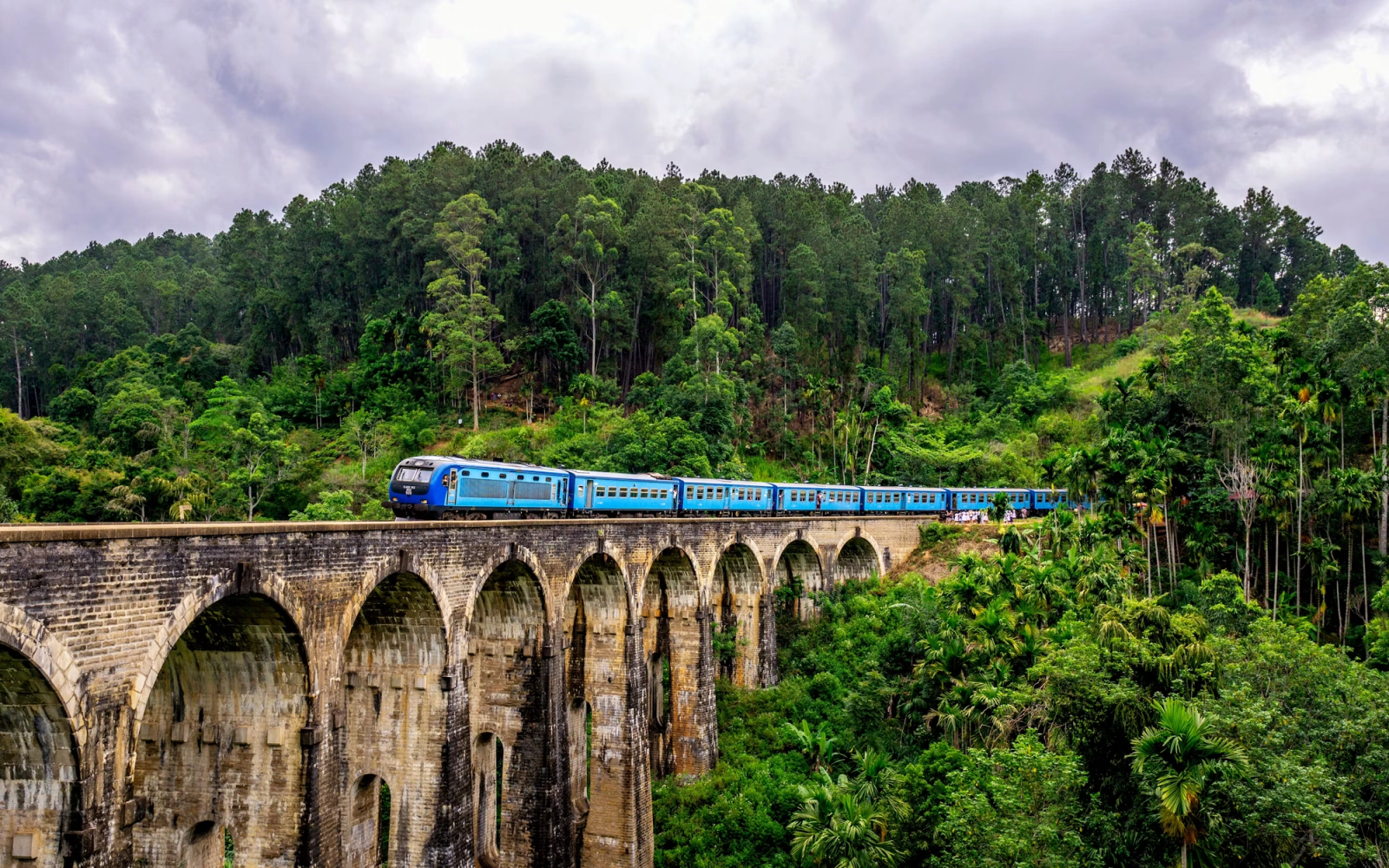 Blue train crossing a stone multi-arched viaduct through forested hills