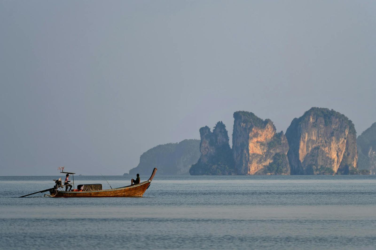 A longtail boat with two passengers crossing calm water near jagged limestone rocks