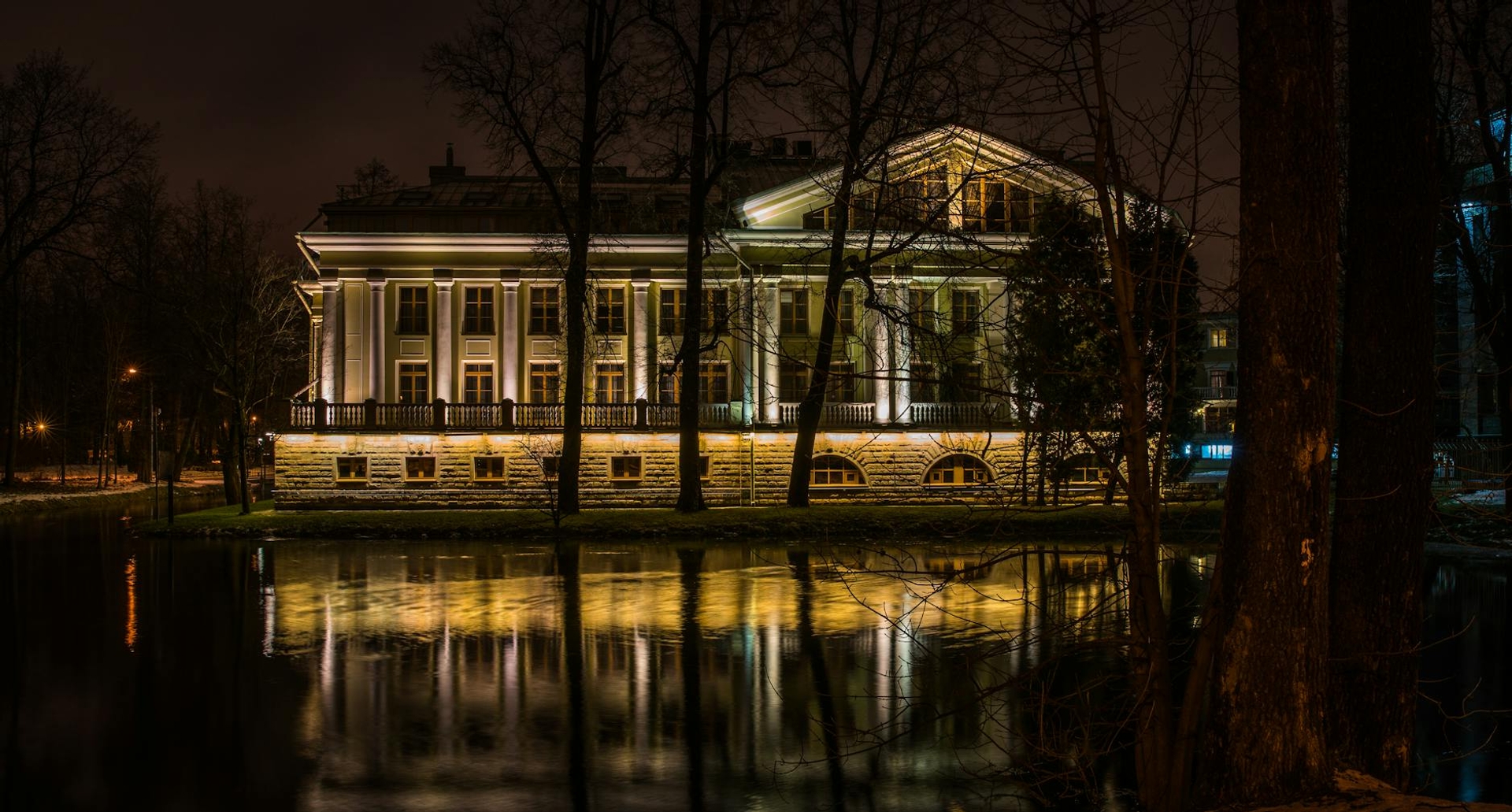 A columned white mansion lit up at night and reflected in still water