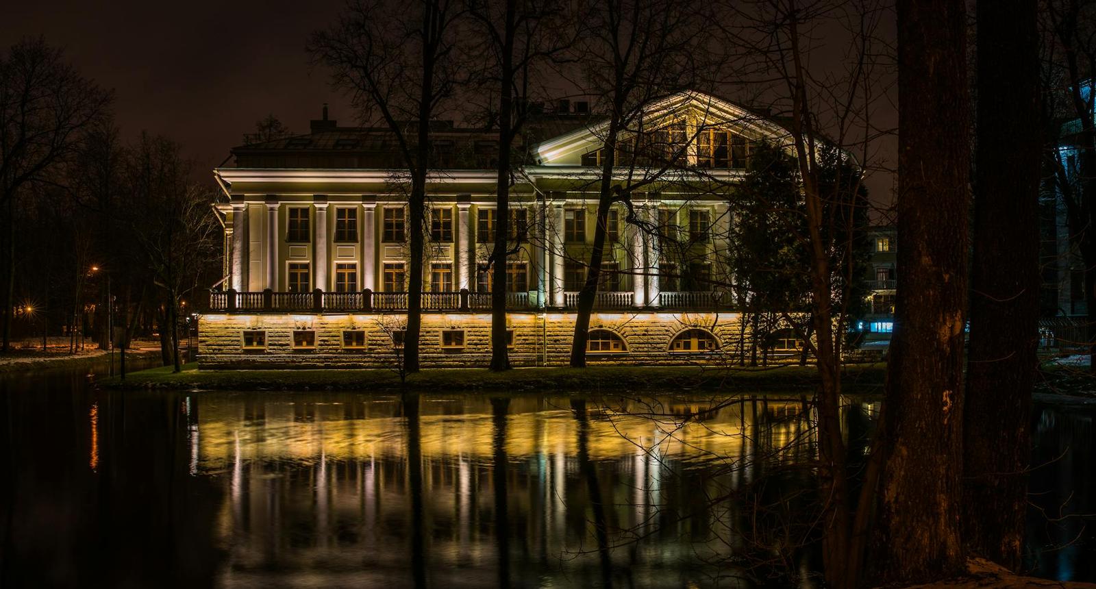 A columned white mansion lit up at night and reflected in still water