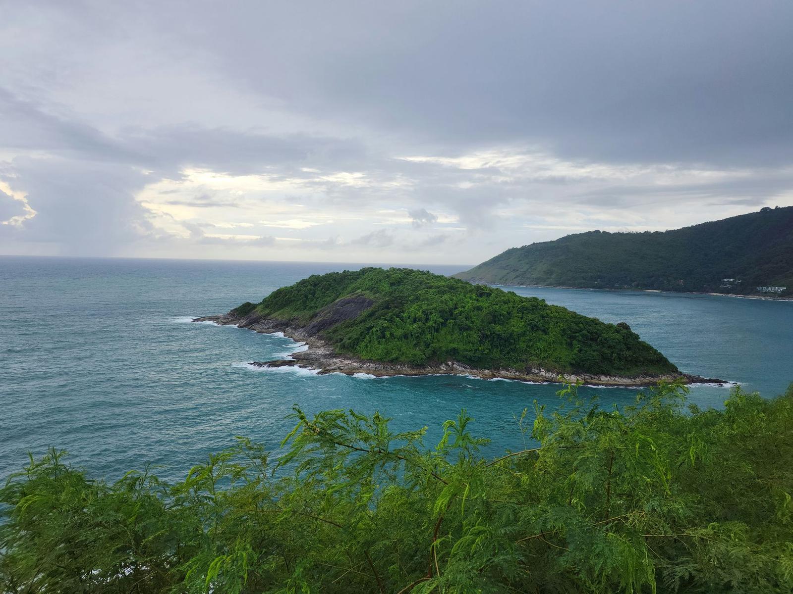 A small forested headland jutting into a blue sea under cloudy sky