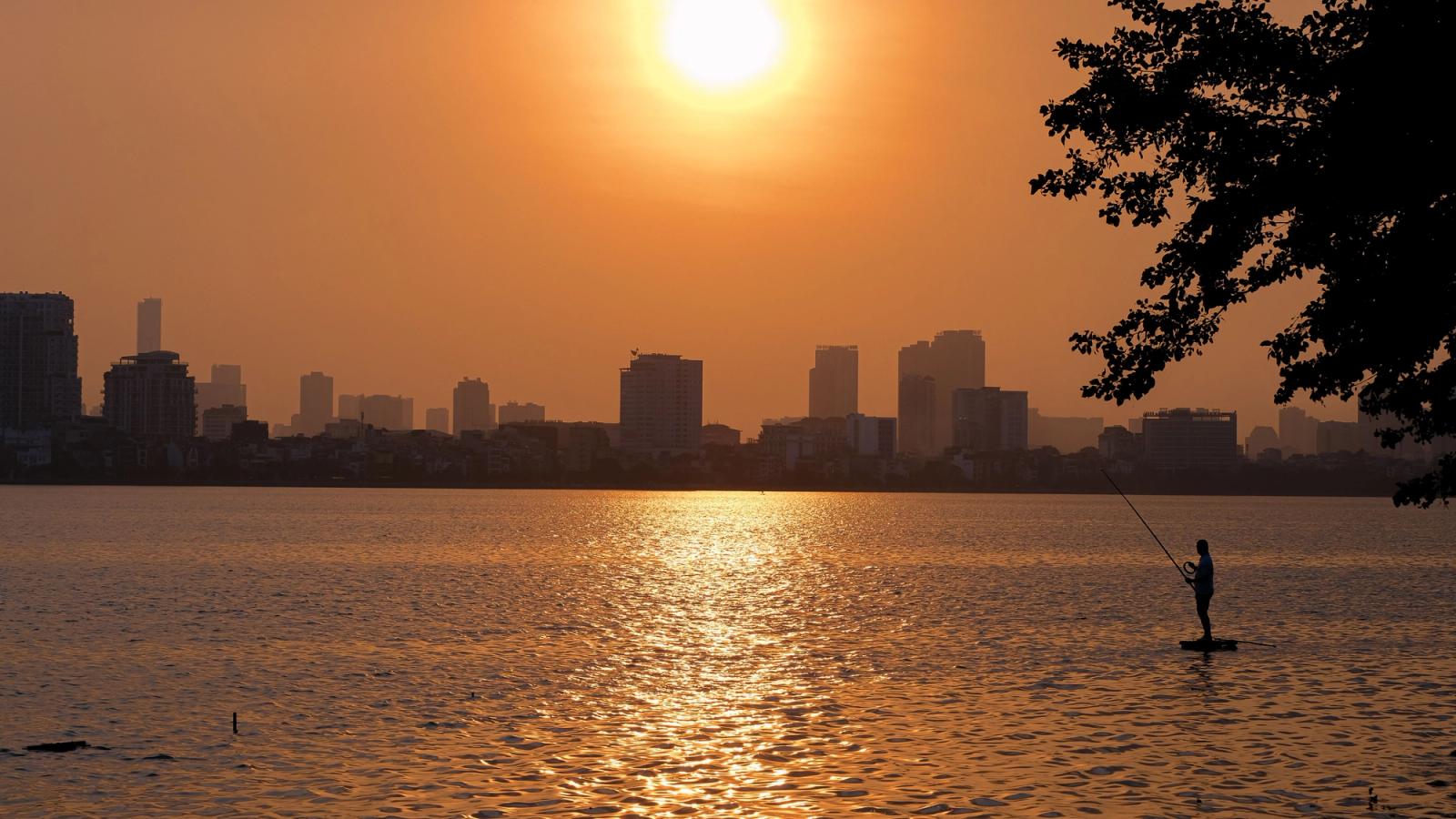 Silhouette of a person fishing at sunset with Hanoi skyline in the background, creating a serene urban scene