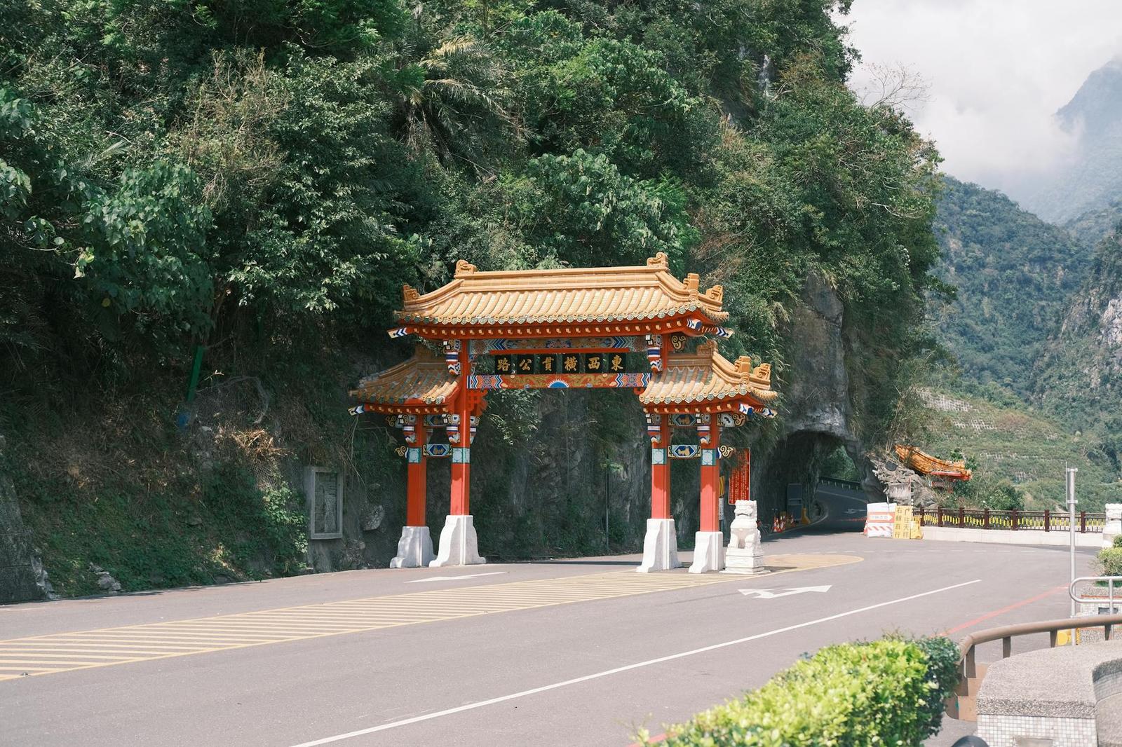 An ornate red and gold Chinese gateway arching over a road at the foot of cliffs