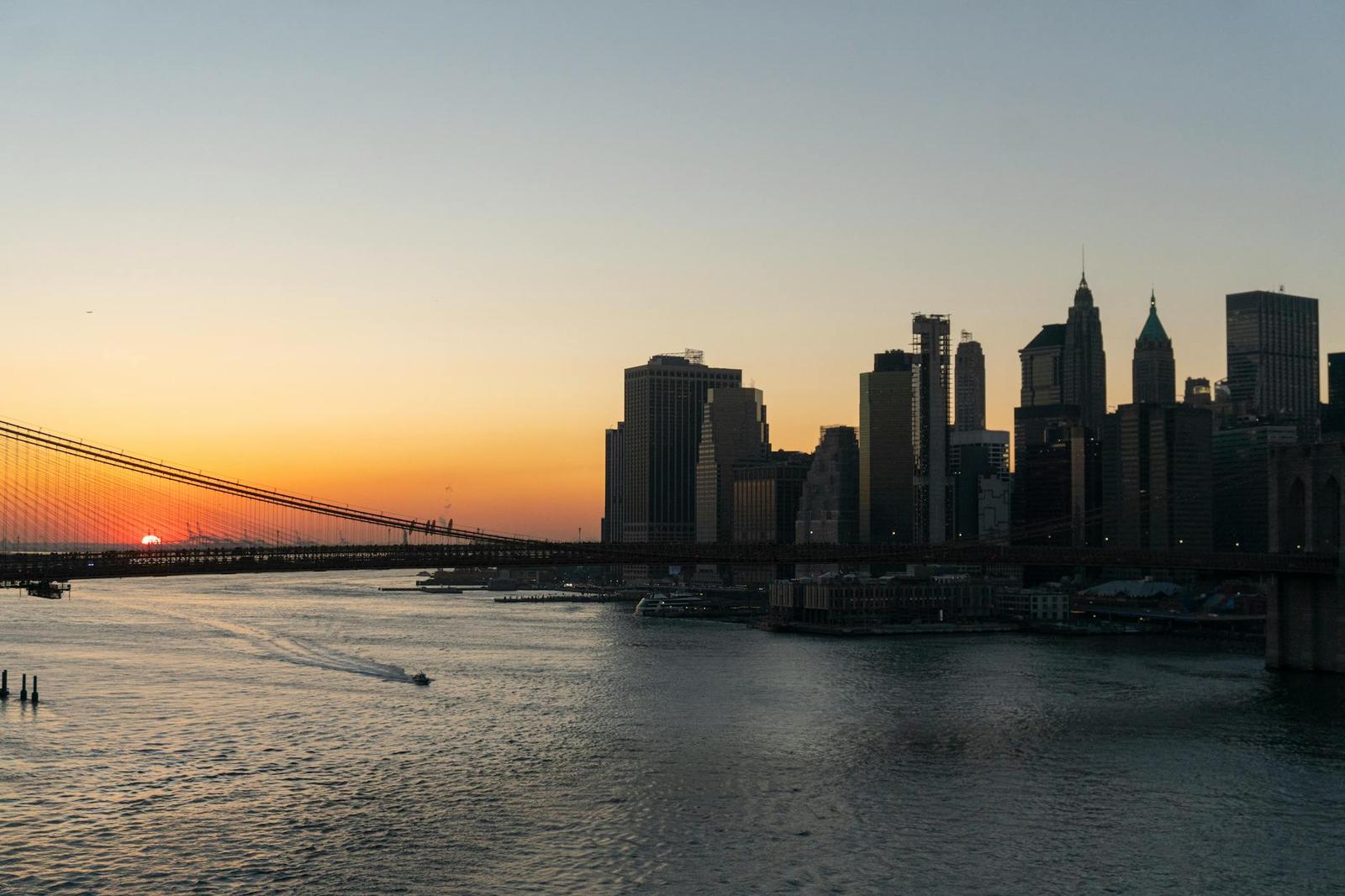 A city skyline and bridge silhouetted against an orange sunset over the water