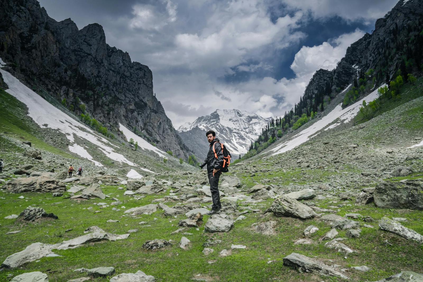 A trekker crossing a green mountain valley flanked by snow patches and peaks