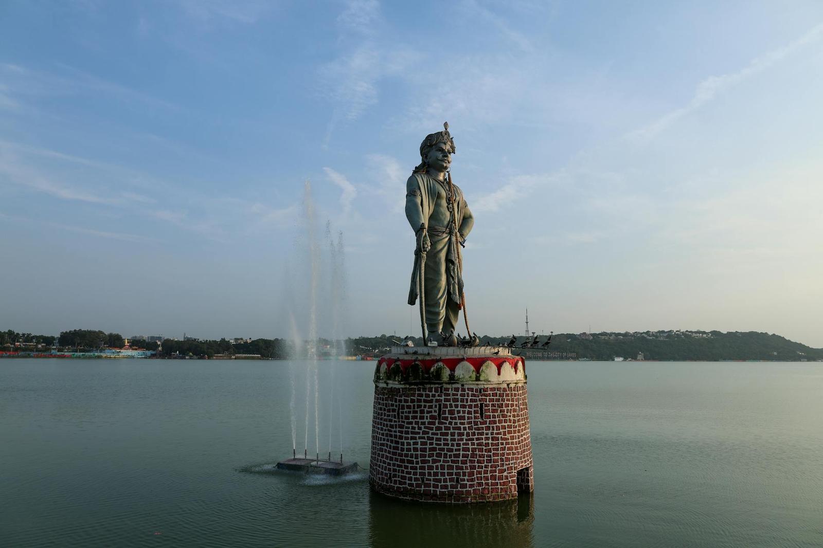 A bronze statue on a stone pedestal rising from a lake beside a fountain