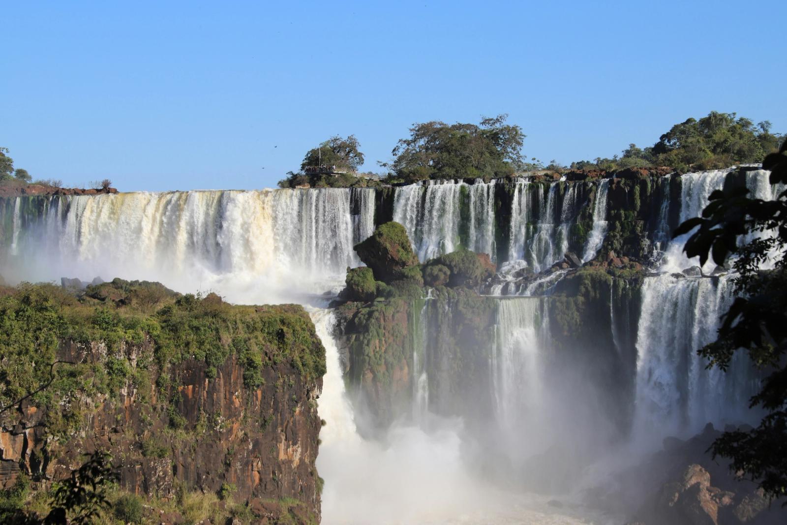 Multiple waterfalls cascade over a cliff into a misty jungle gorge
