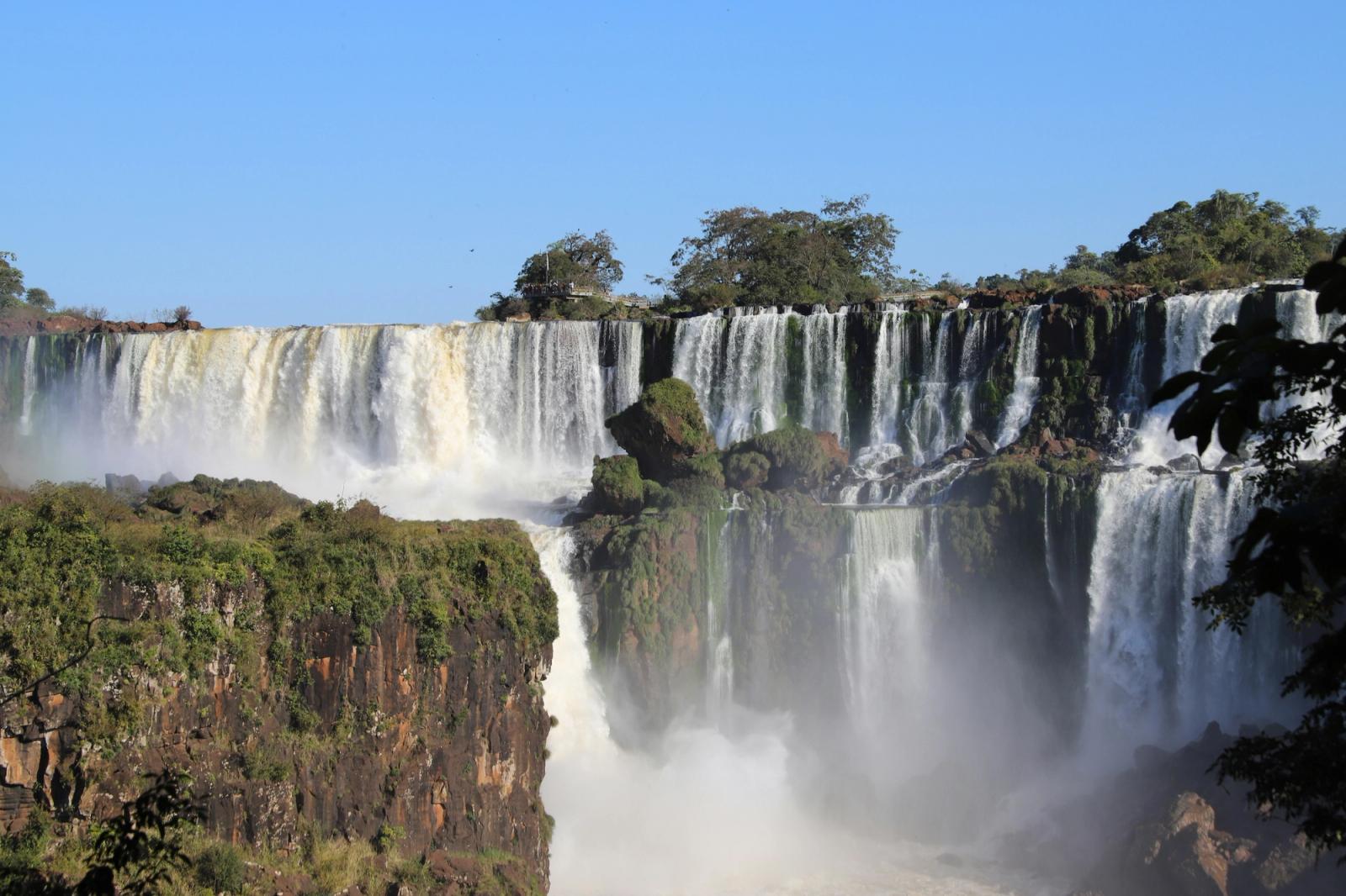 Multiple waterfalls cascade over a cliff into a misty jungle gorge