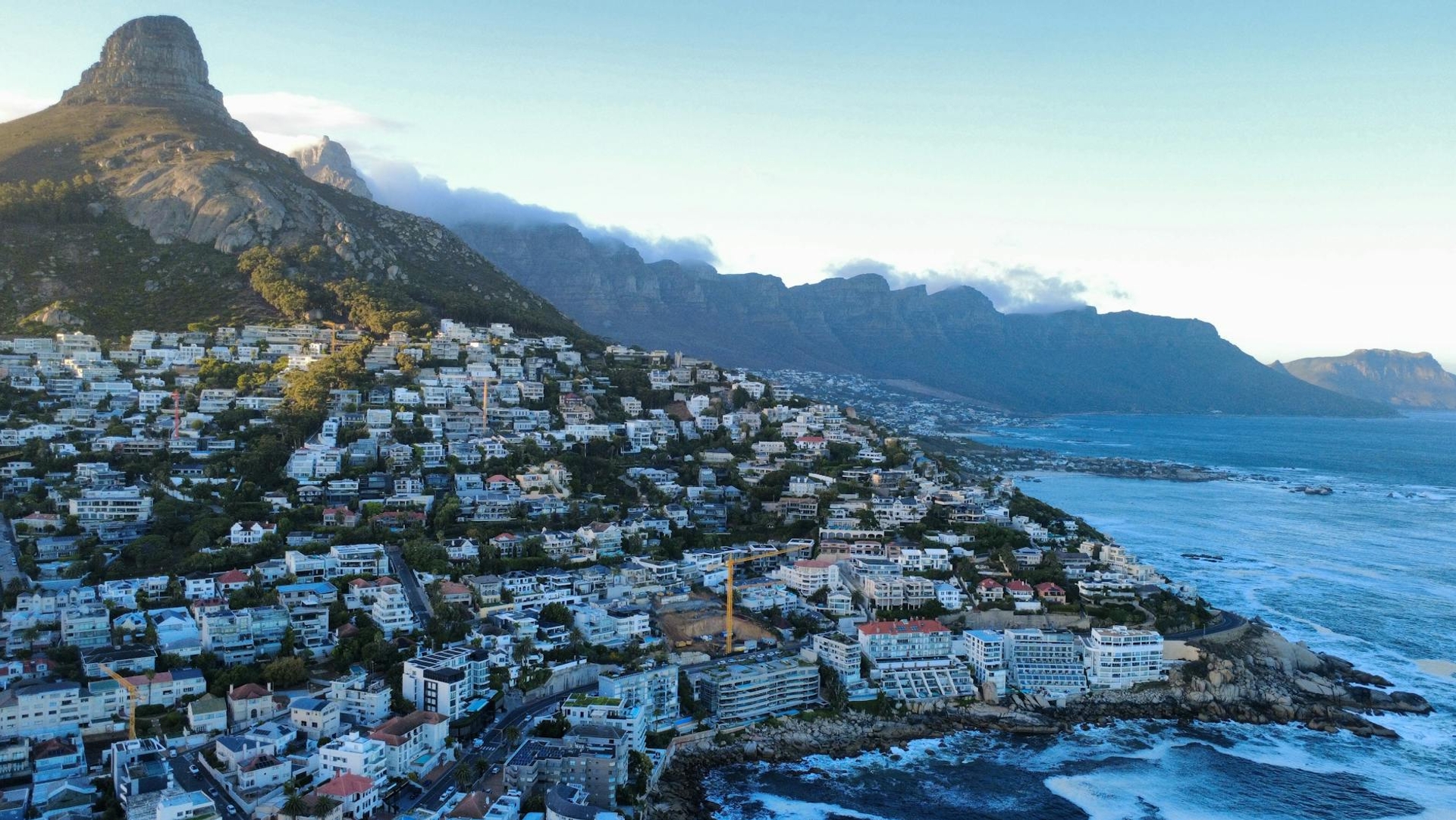 A coastal city spreads below a rocky peak and cloud-draped mountains at dusk