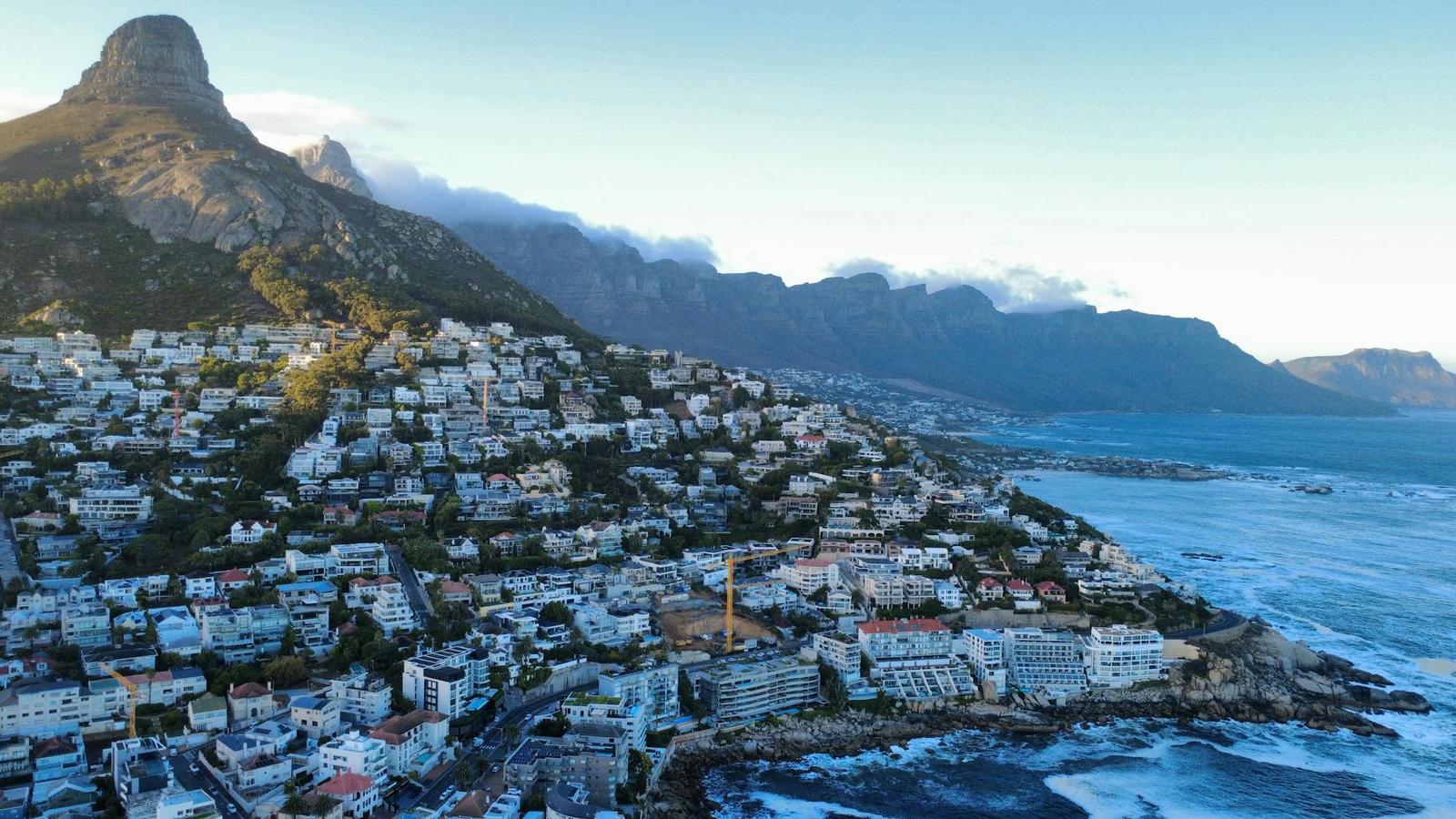 A coastal city spreads below a rocky peak and cloud-draped mountains at dusk