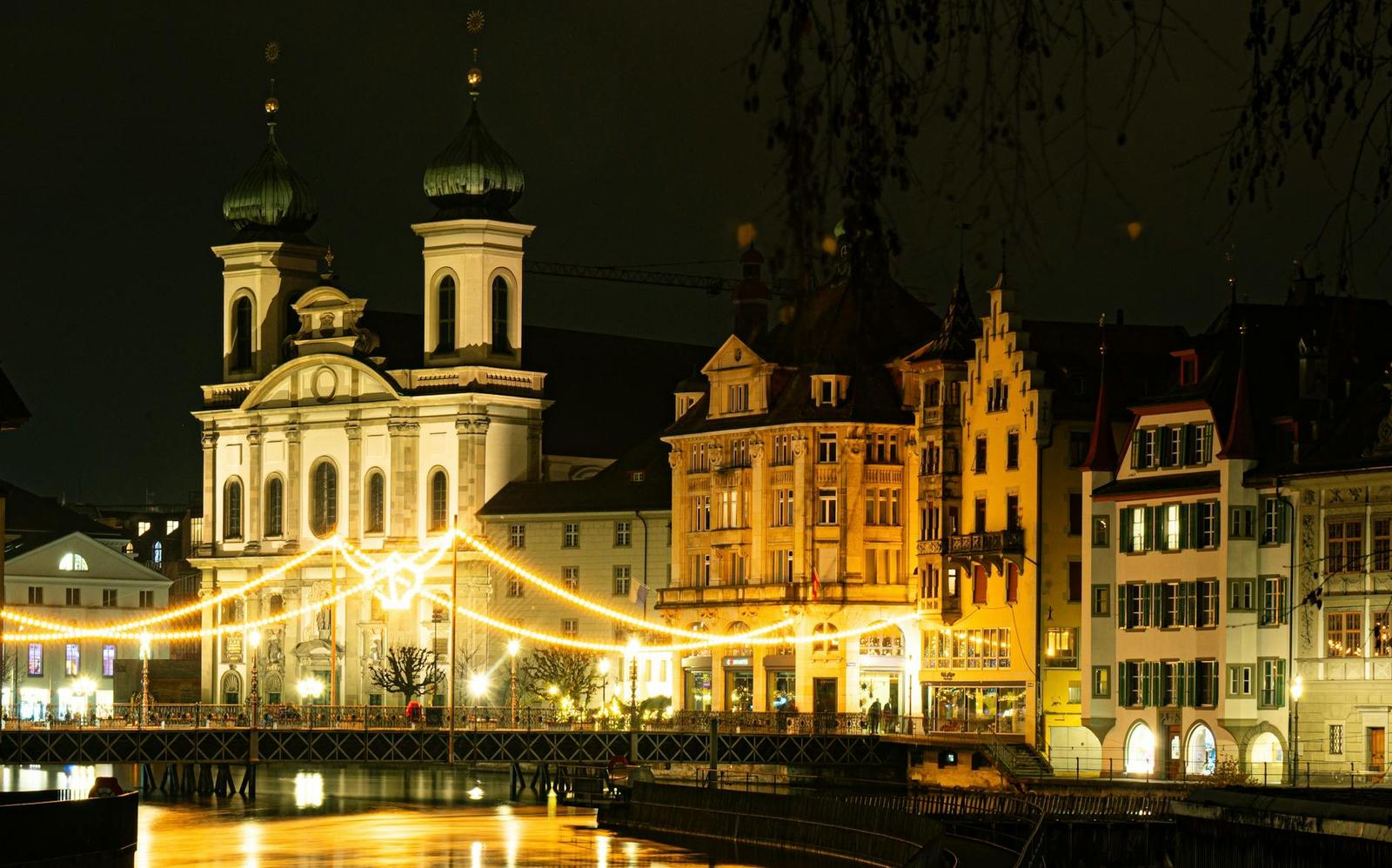 A lit baroque church and old-town buildings beside a river strung with festive lights