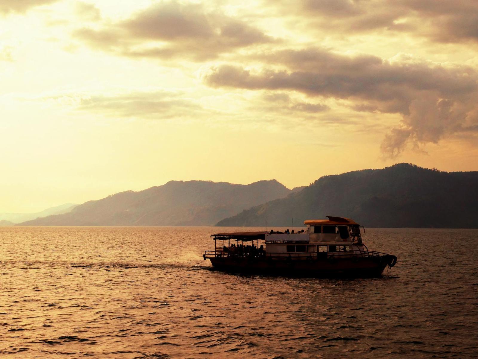 A tour boat crosses calm water at golden sunset with hills silhouetted behind