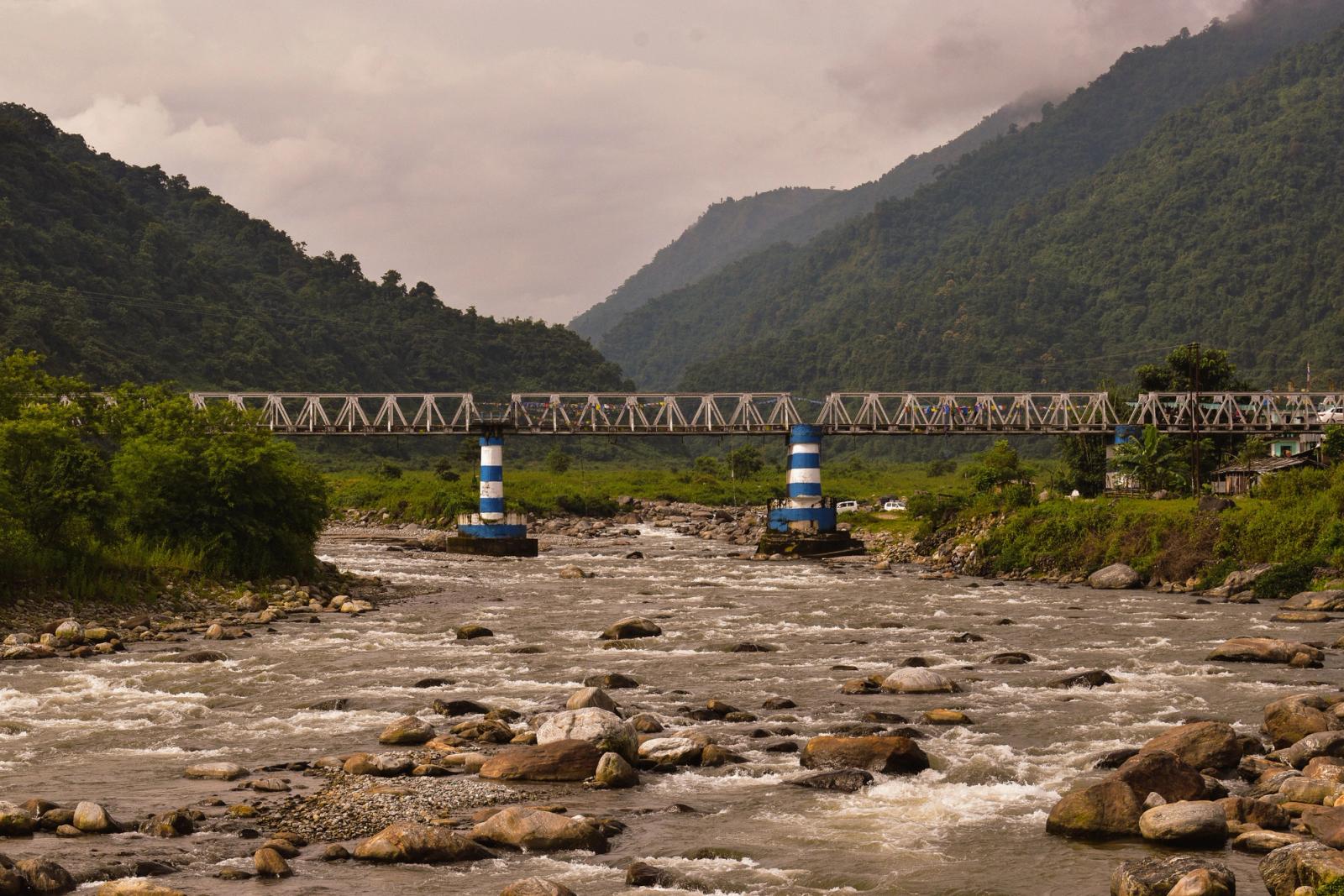 A picturesque view of a river flowing under a steel bridge in India, surrounded by lush mountains