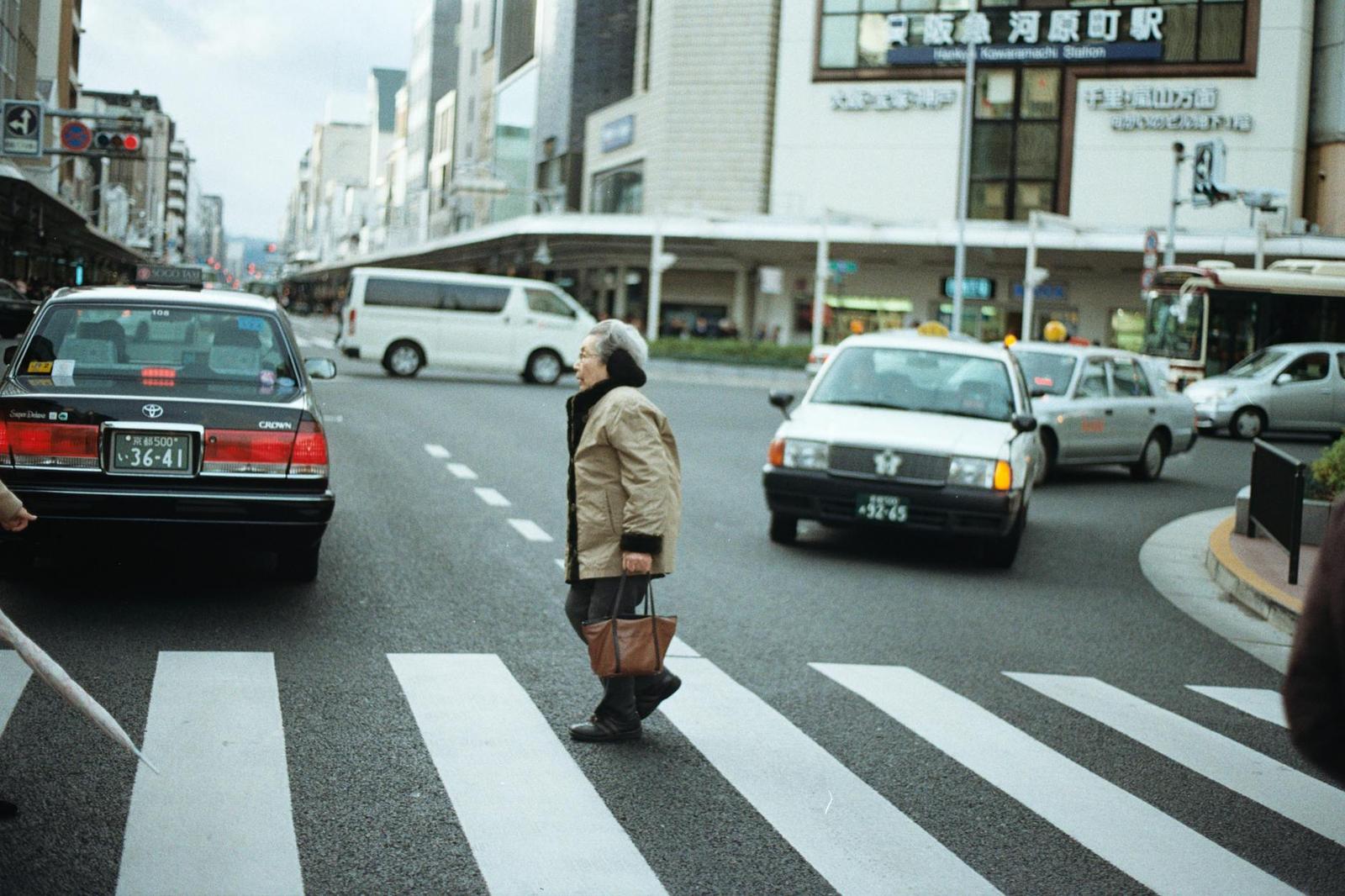 An elderly woman crossing a city crosswalk between waiting cars