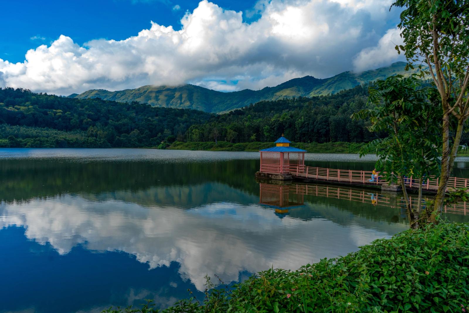 Tranquil lake scene in HirekoLale, India, with a dock, lush greenery, and mountain backdrop under blue skies and fluffy clouds