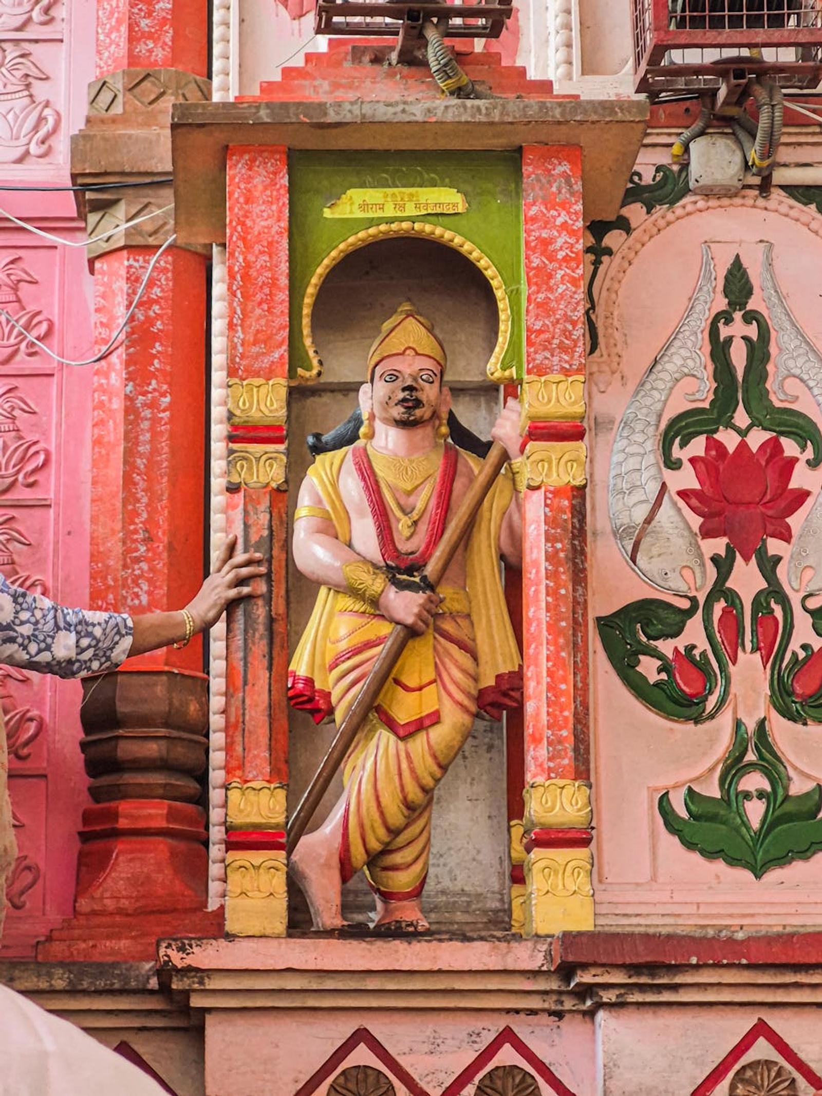 Brightly painted deity statue in an ornate temple niche with a devotee's hand resting on the frame