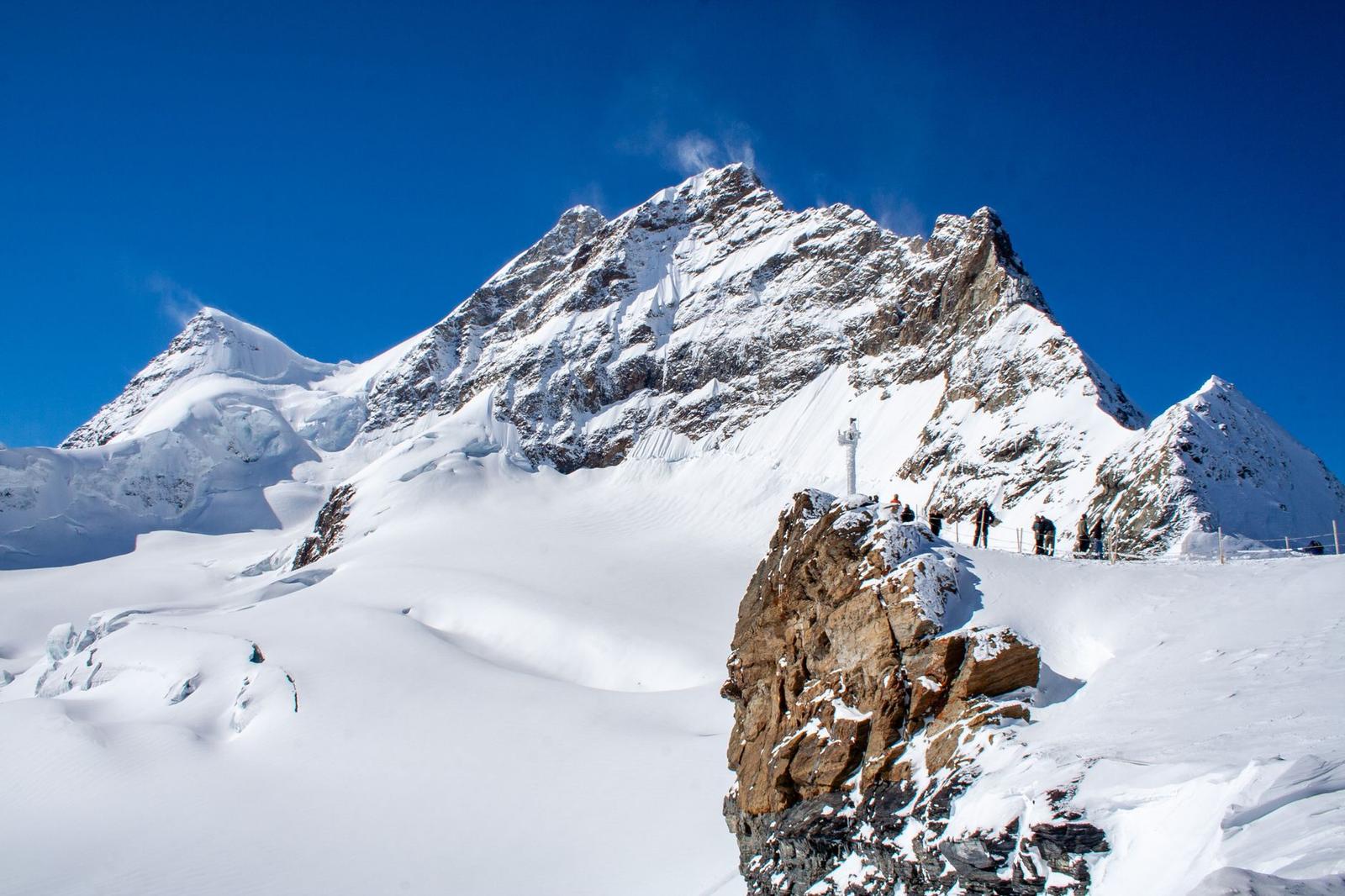 Hikers crossing a snowy ridge below a jagged mountain peak under blue sky