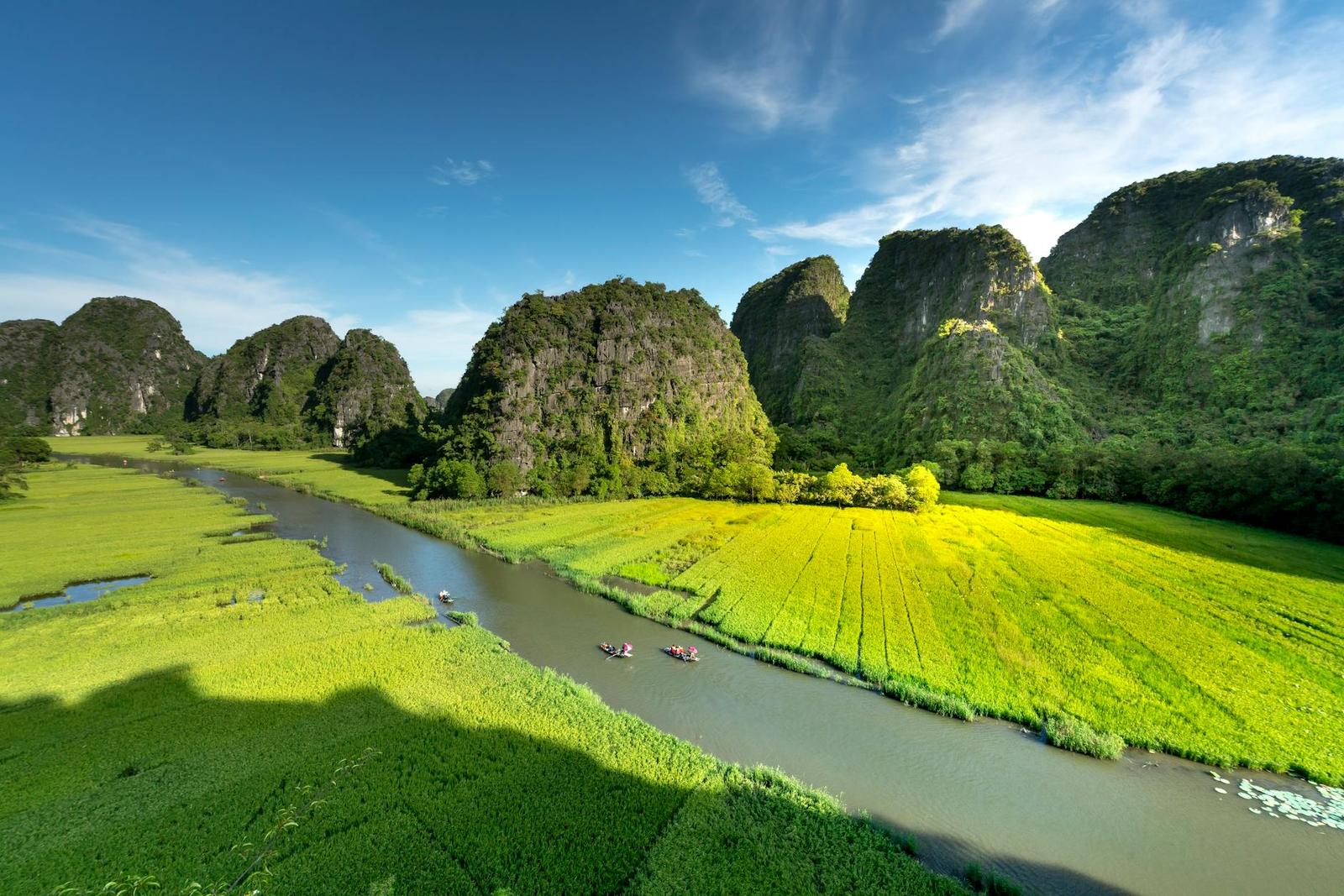 Aerial view of a scenic river cutting through lush green fields with towering limestone mountains under a clear sky