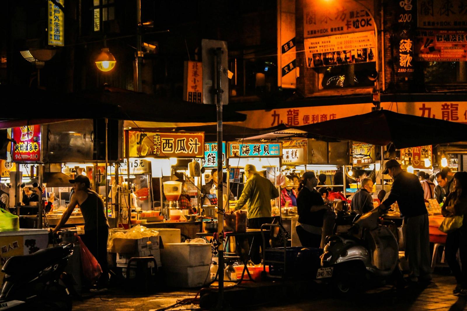 A bustling night market street lined with stalls, signage and parked scooters