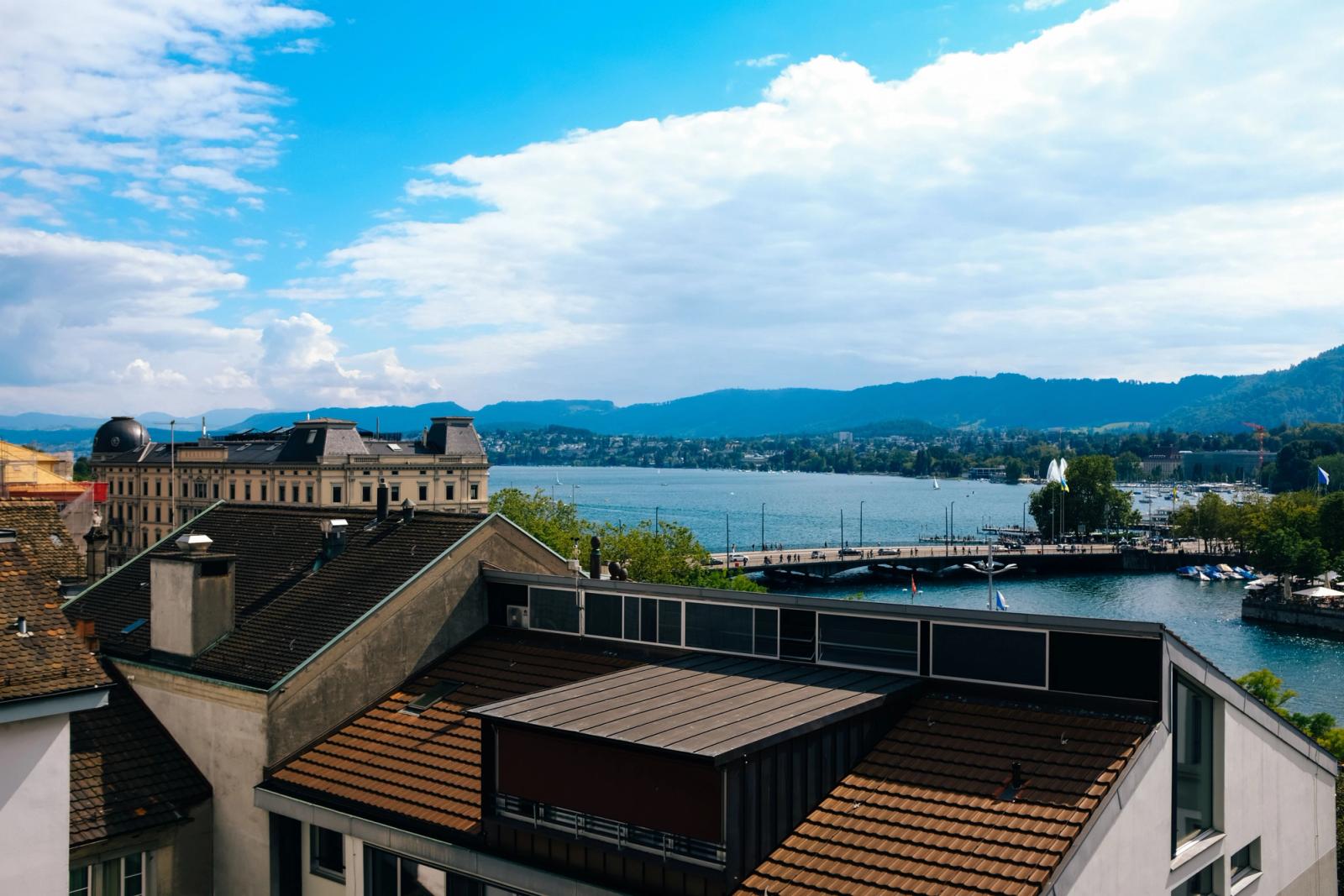 Panoramic view of Zurich featuring the Limmat River, rooftops, and mountains under a bright blue sky