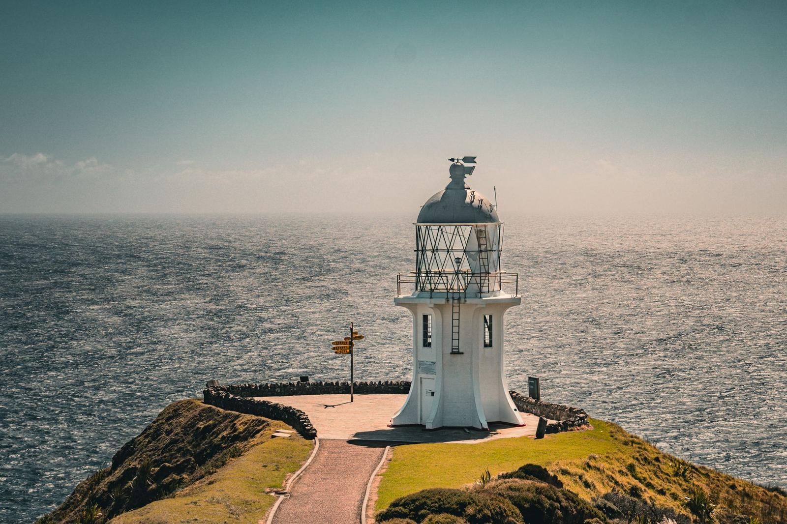A white lighthouse on a grassy headland above a hazy ocean horizon
