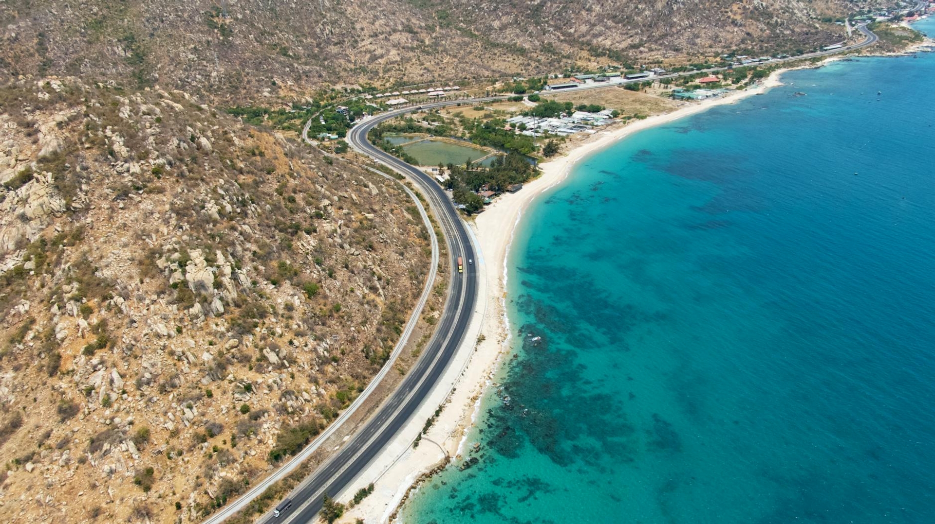 Coastal road winding along a dry hillside beside a turquoise bay