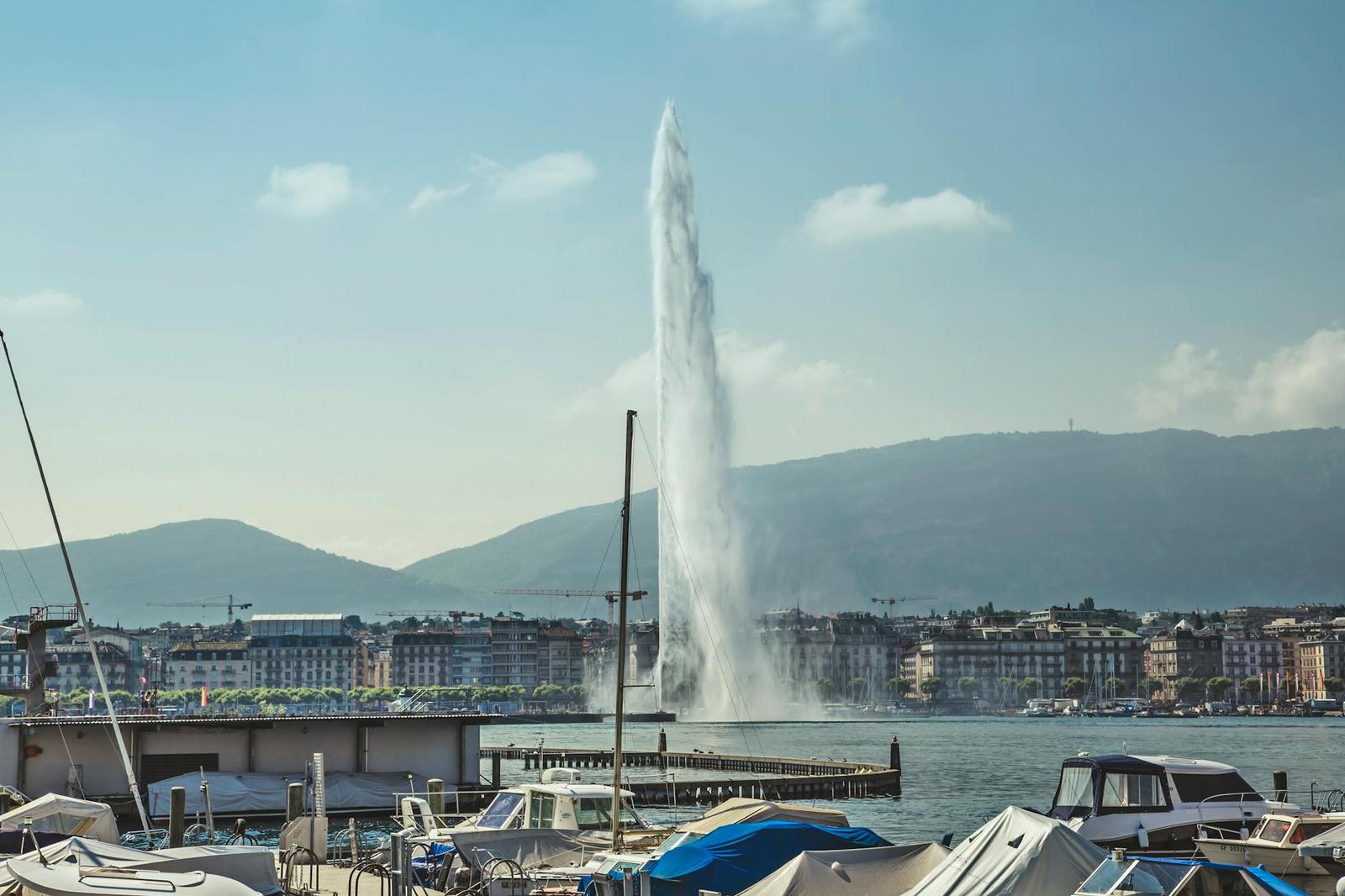 Lakeside fountain seen past moored boats and a marina at sunset