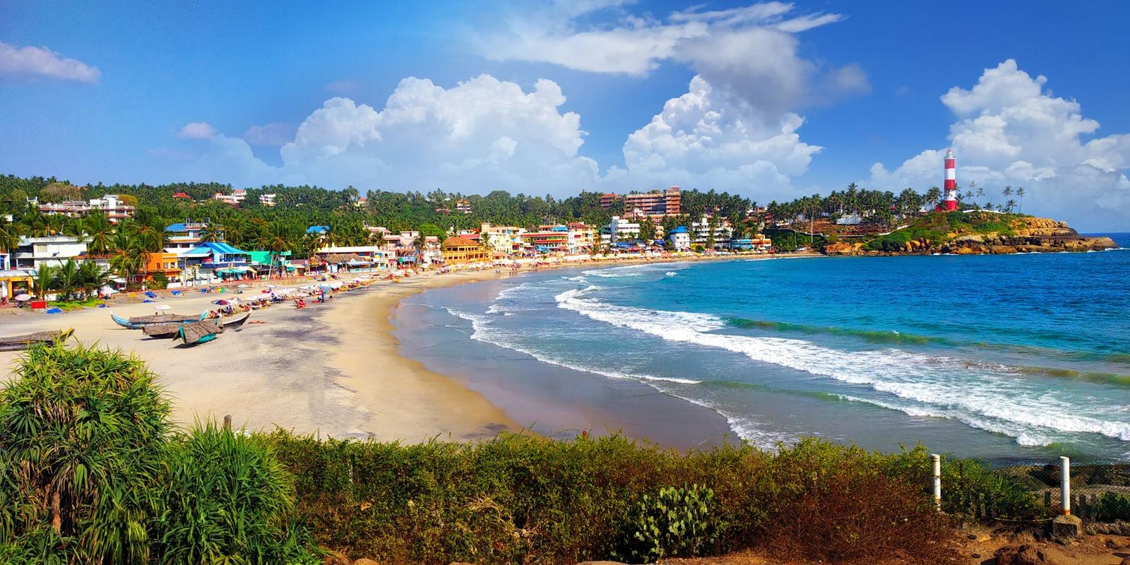 A curved sandy beach lined with palm-fringed buildings and a striped lighthouse