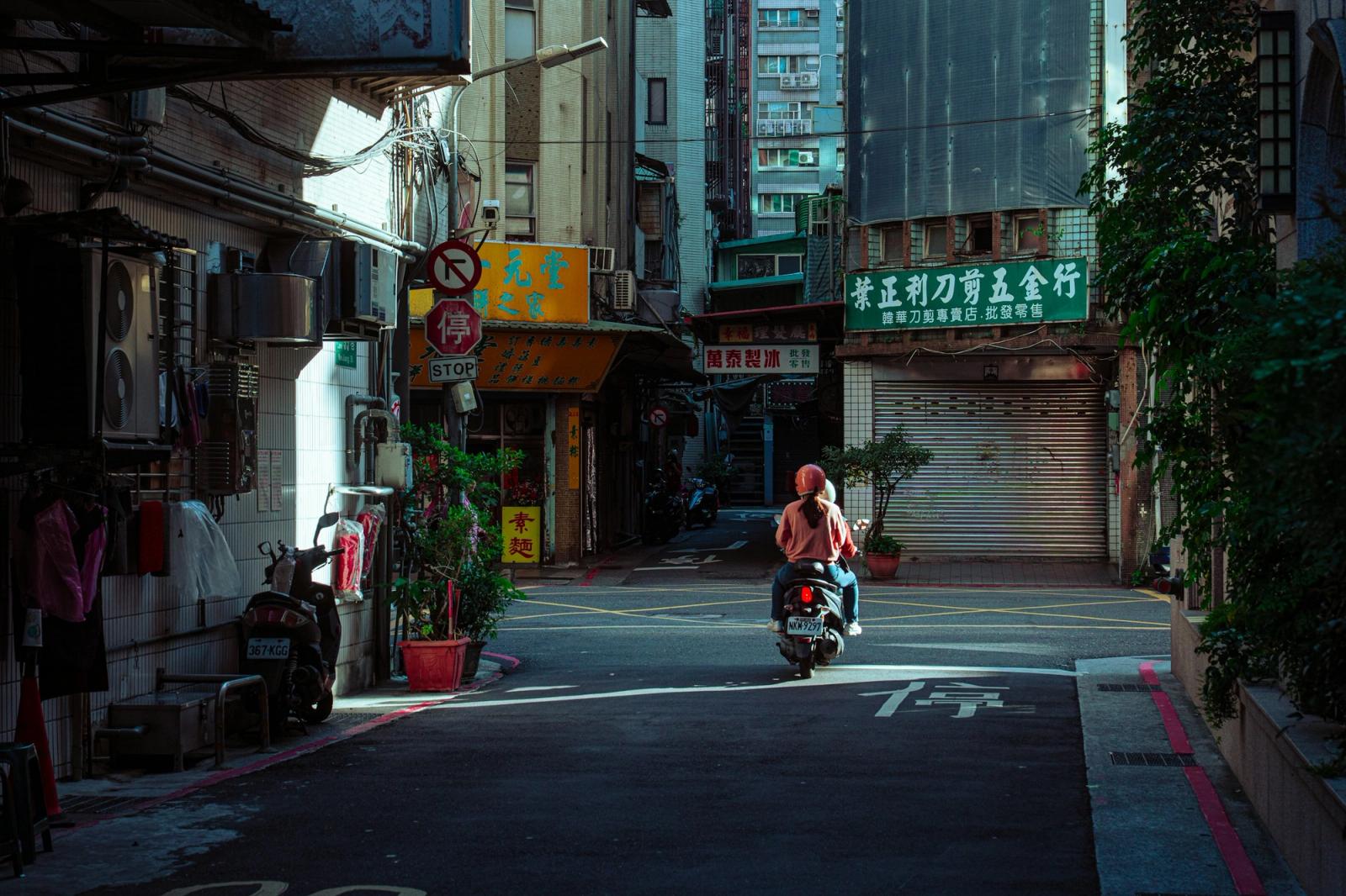 A rider on a scooter passes through a narrow alley lined with shopfronts and signage