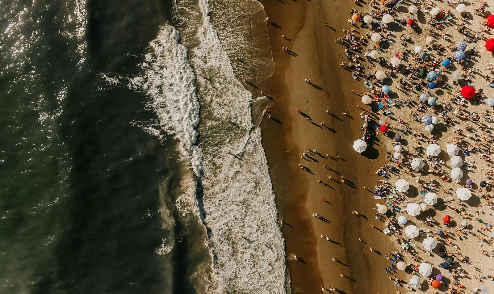 Aerial view of a crowded beach with colourful umbrellas beside dark waves