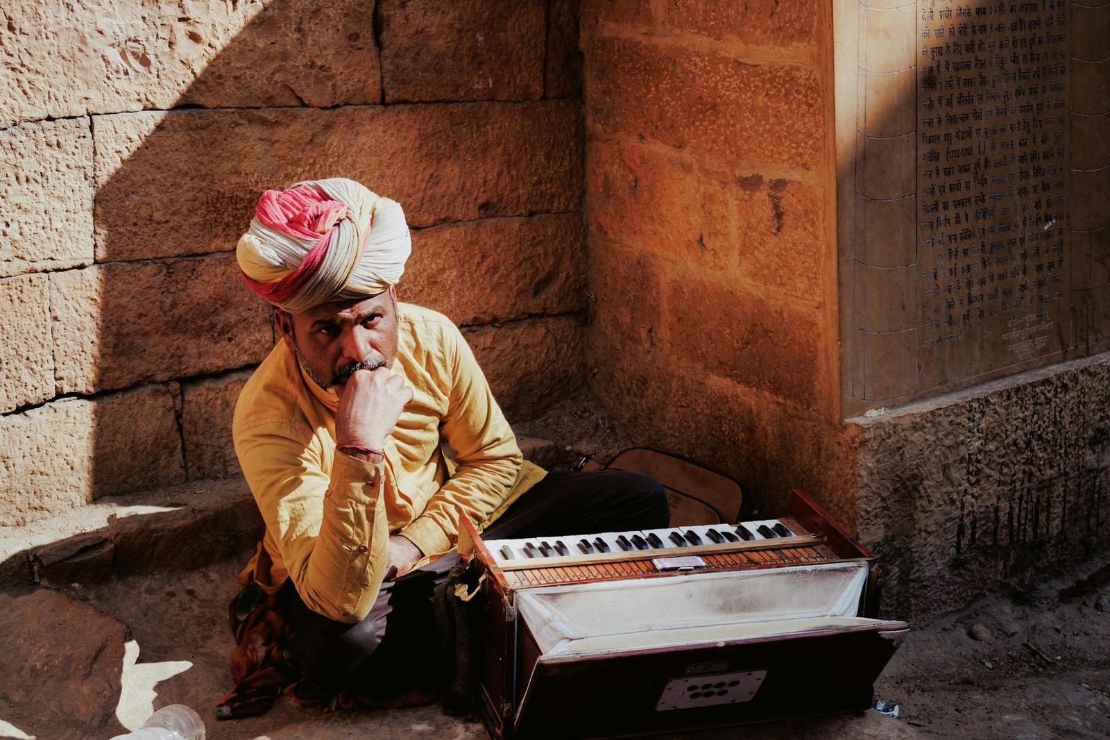 A turbaned musician seated against a stone wall with a harmonium